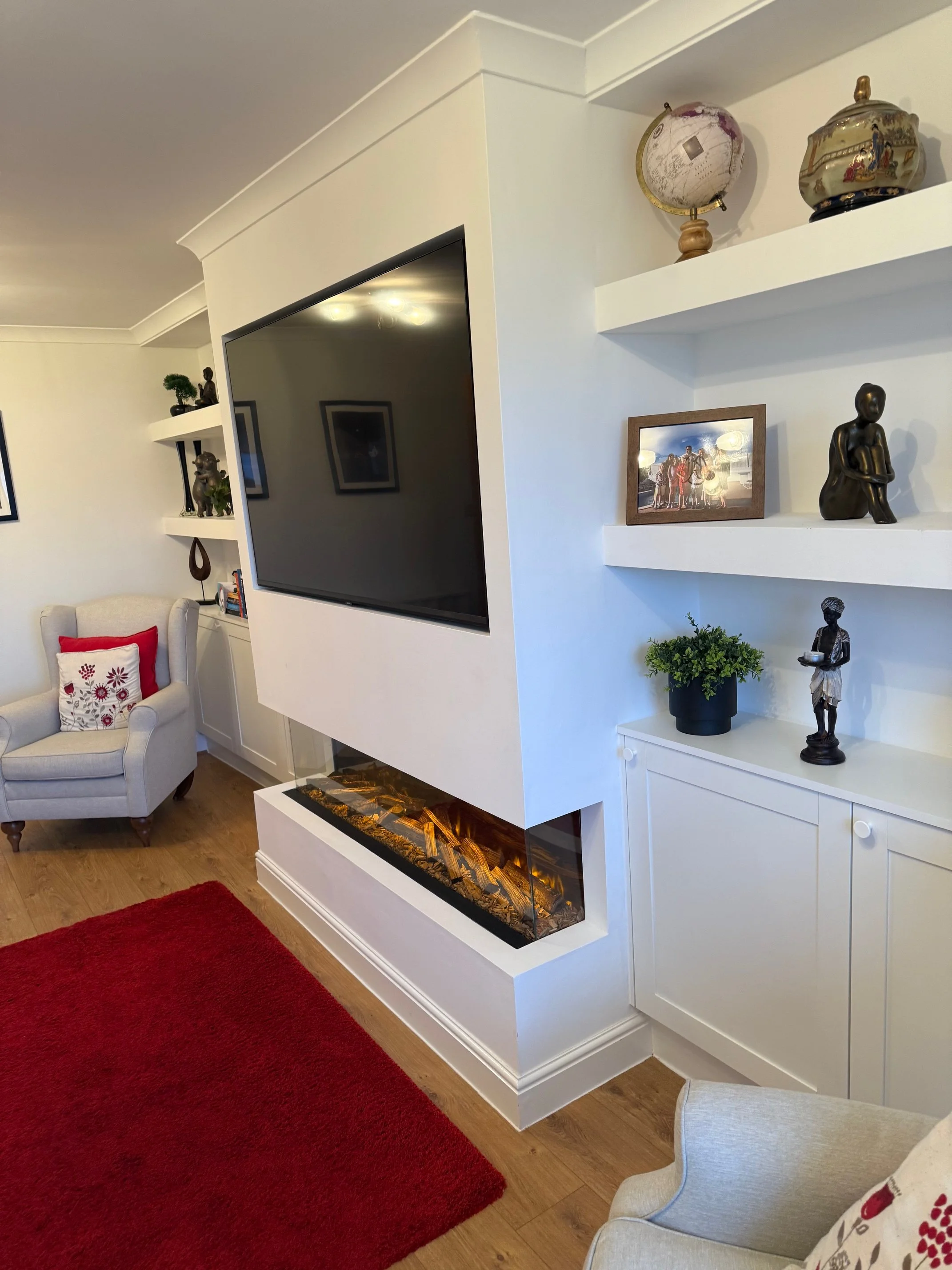 Living room with a wall-mounted flat screen TV, modern electric fireplace, white shelving with decorative items, framed family photo, potted plant, and sculpture. Beige armchair with red pillow, red rug, and wooden flooring.