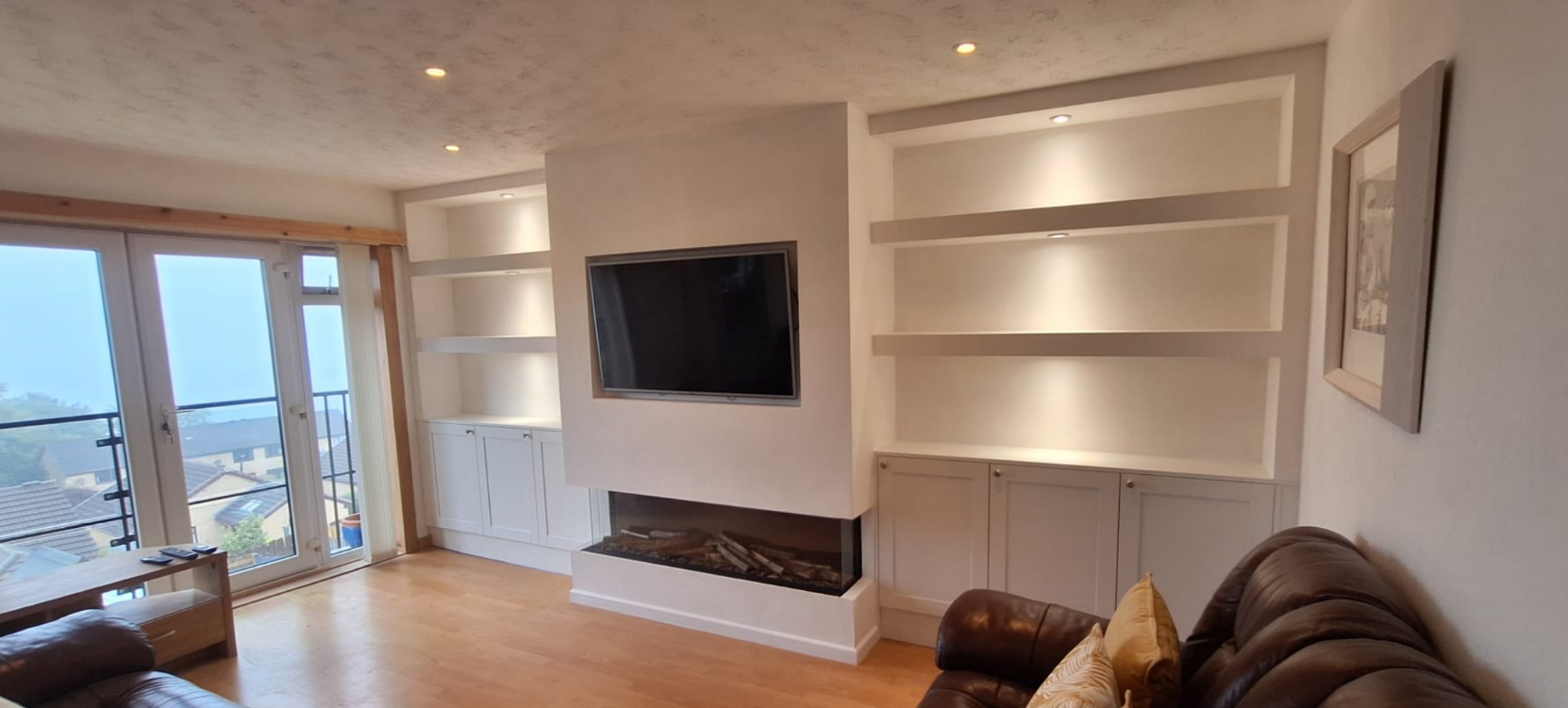 Living room with sliding glass doors leading to a balcony, wooden flooring, a wall-mounted TV above a fireplace, built-in white shelves, a leather sofa with yellow pillows, and a framed picture on the wall.