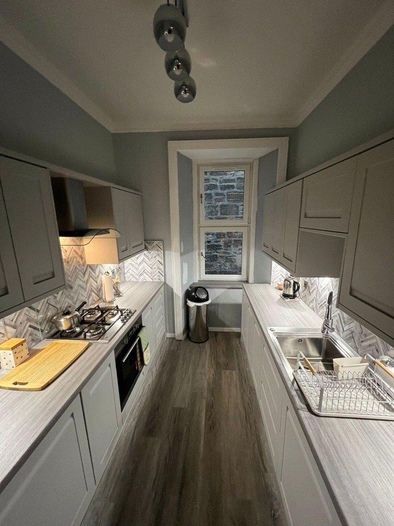 Small modern galley kitchen with light gray cabinets, herringbone tile backsplash, gray countertops, stainless steel appliances, and a window at the end.