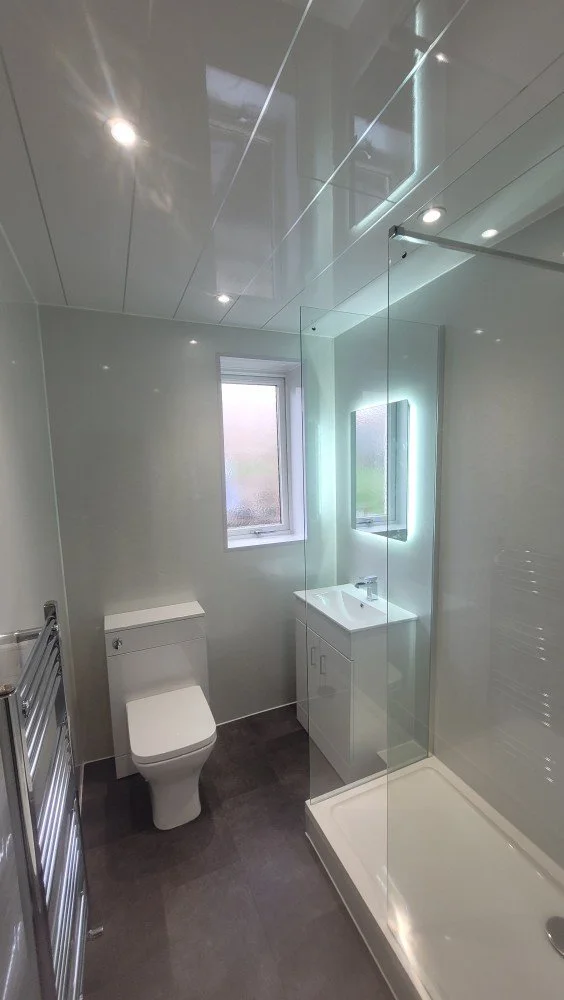 Modern small bathroom with a toilet, a white vanity with a mirror and light, a window, and a glass shower enclosure with a white shower tray.