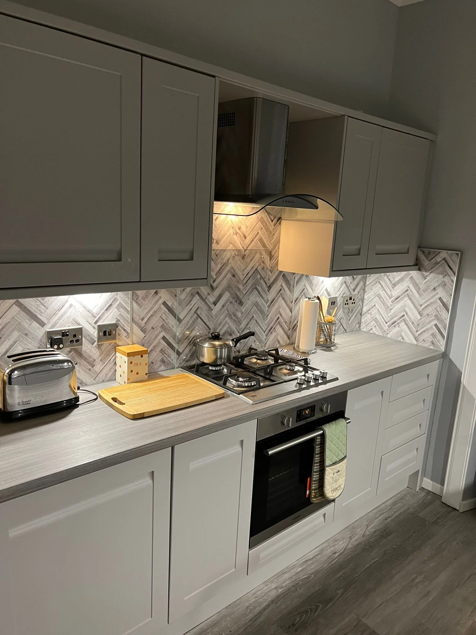 Modern kitchen with gray cabinets, a gray countertop, a built-in oven, a gas stove with a kettle on it, a toaster, a paper towel roll, and a textured tile backsplash.