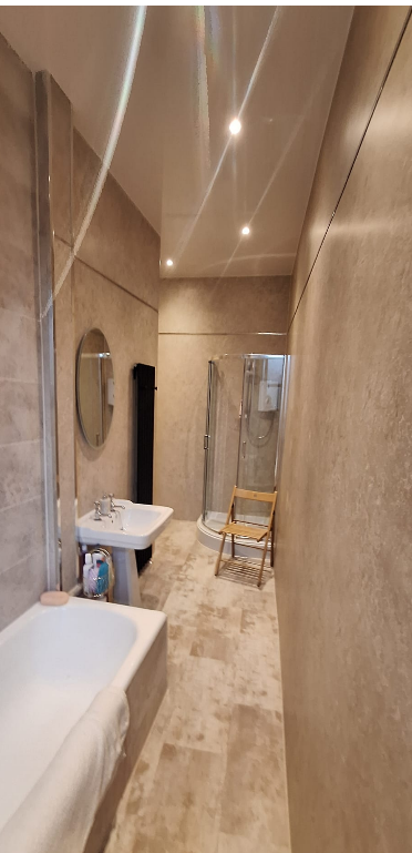 Narrow bathroom with beige walls and floor, featuring a small sink, round mirror, shower enclosure, a wooden chair, and minimal decor.