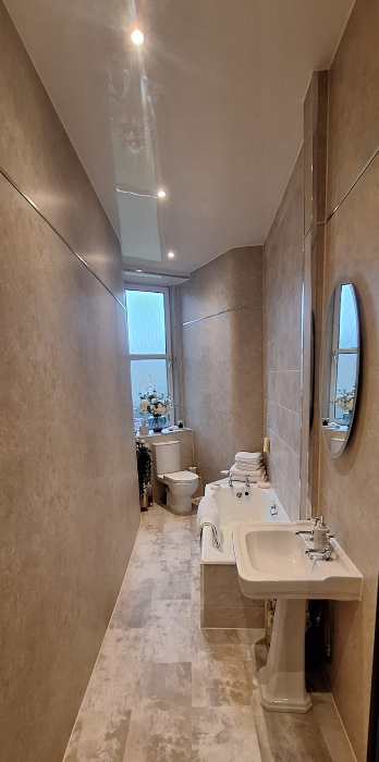 Long, narrow bathroom with a window, toilet, bathtub, pedestal sink, mirror, and beige tiled walls and floors.