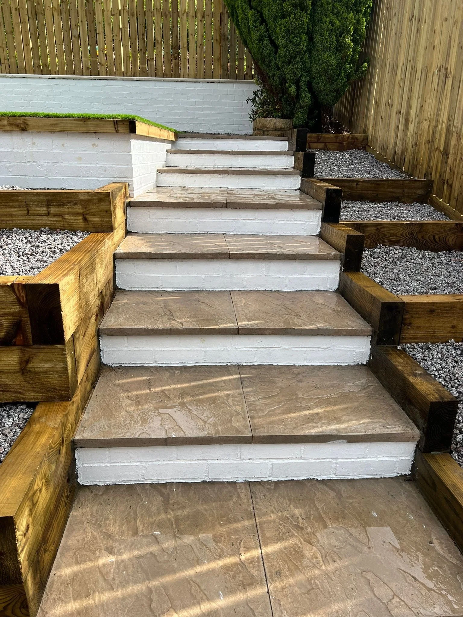Wooden and concrete stairs leading up to a yard with a white brick wall and a large green bush, surrounded by wooden fencing and retaining walls.