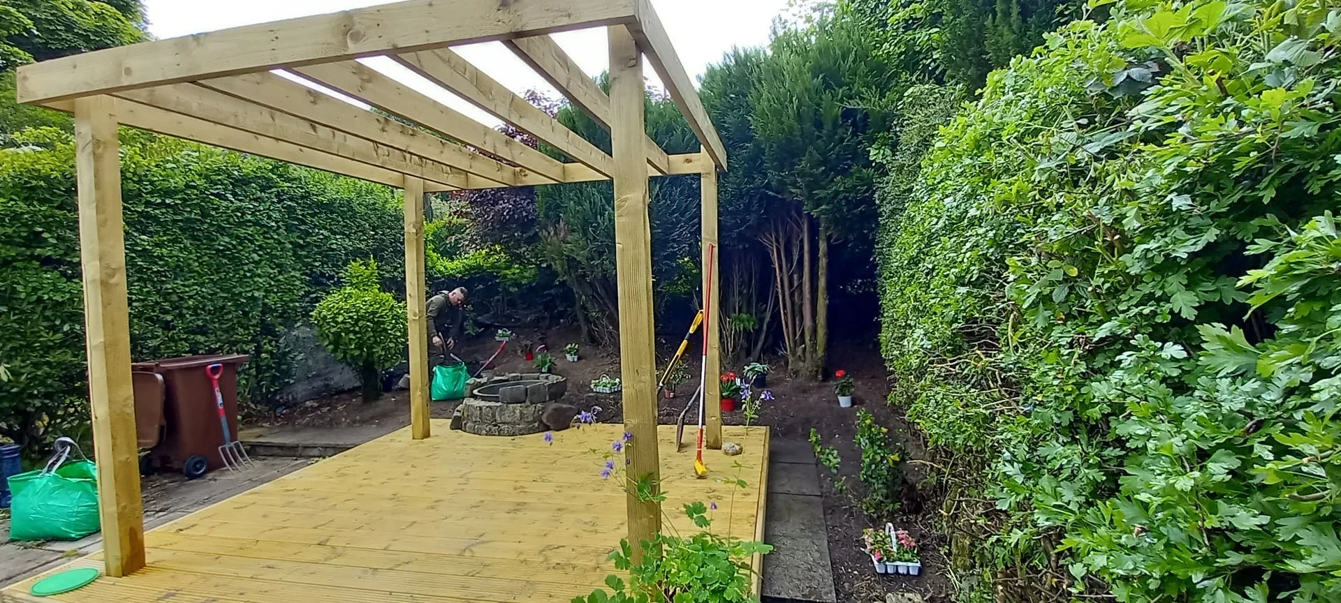 A backyard garden with a wooden deck under construction, surrounded by green bushes and trees, with a person working in the background.