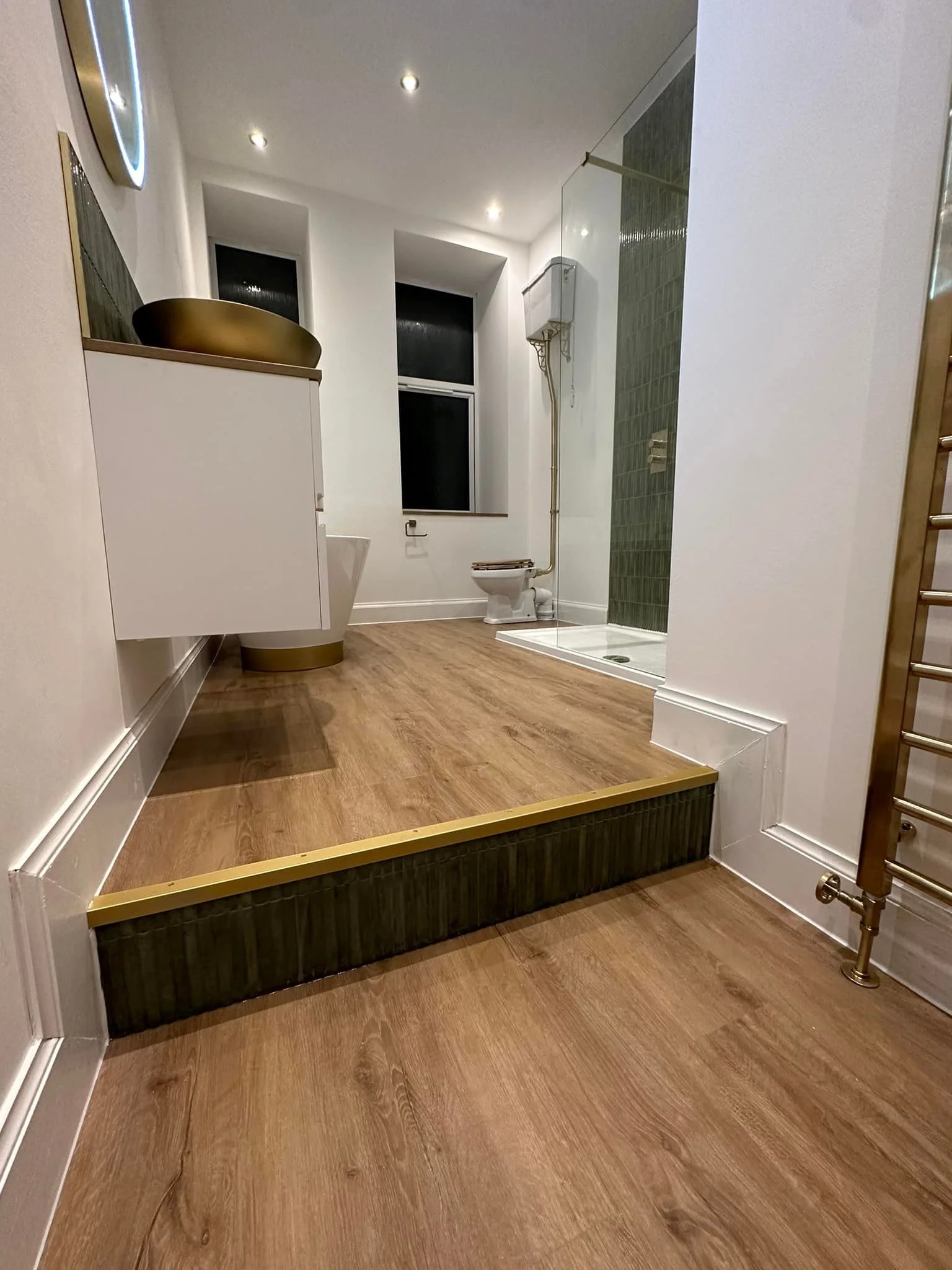 Modern bathroom with wooden floor, white walls, and a raised shower area with dark green tiles. Features a toilet, a white cabinet with a gold sink on top, a window, and a heated towel rail.