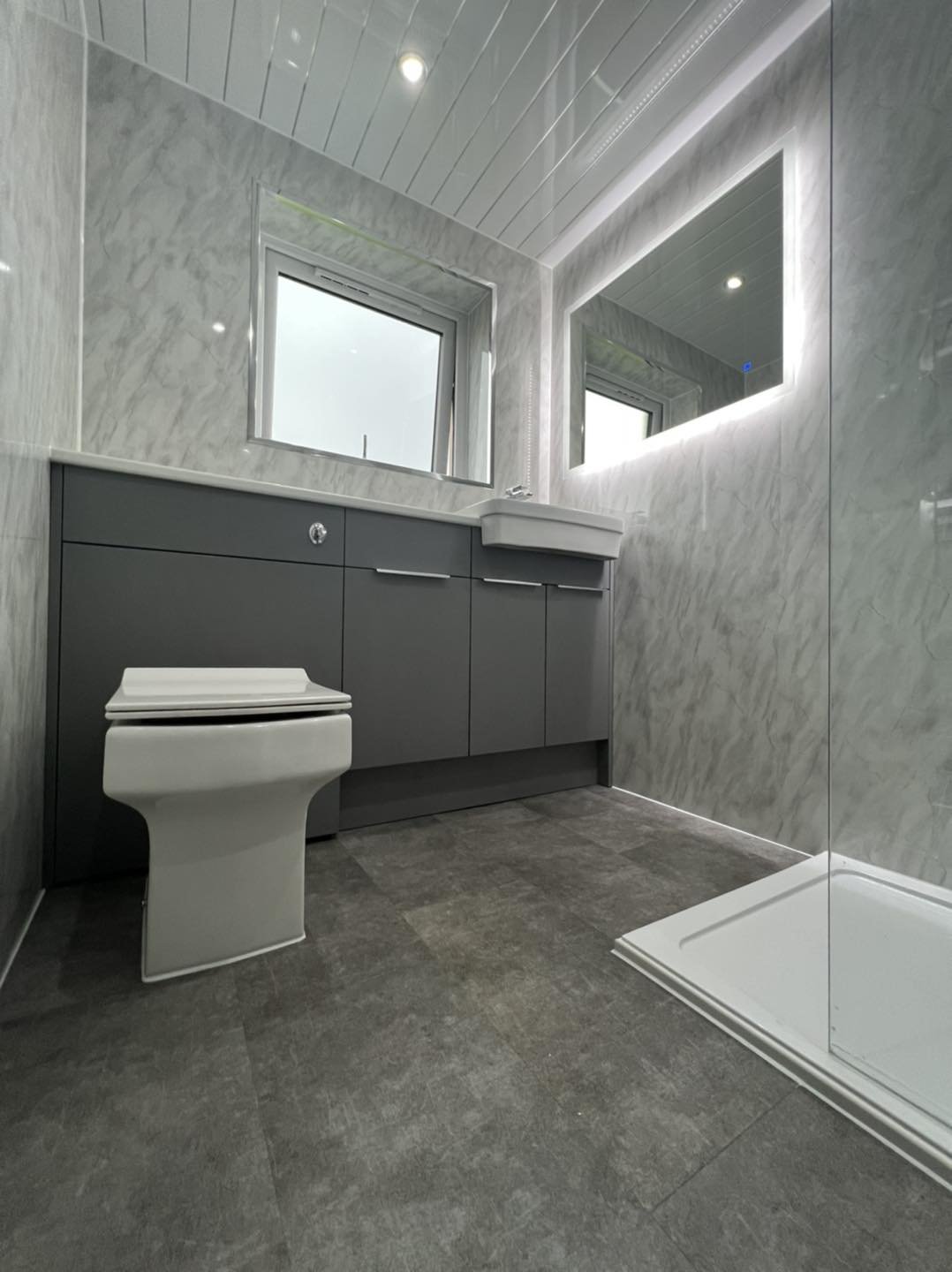 Modern bathroom with gray cabinetry, a white toilet, a small sink, a large mirror with LED lighting, two windows, and a shower area with a white shower base.