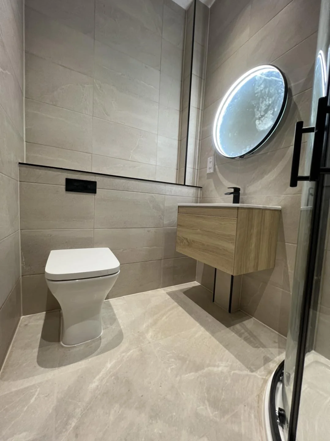Modern bathroom with beige tiled walls and floor, a white toilet, a wall-mounted wooden vanity with a black faucet, a round backlit mirror, and a corner glass shower enclosure.