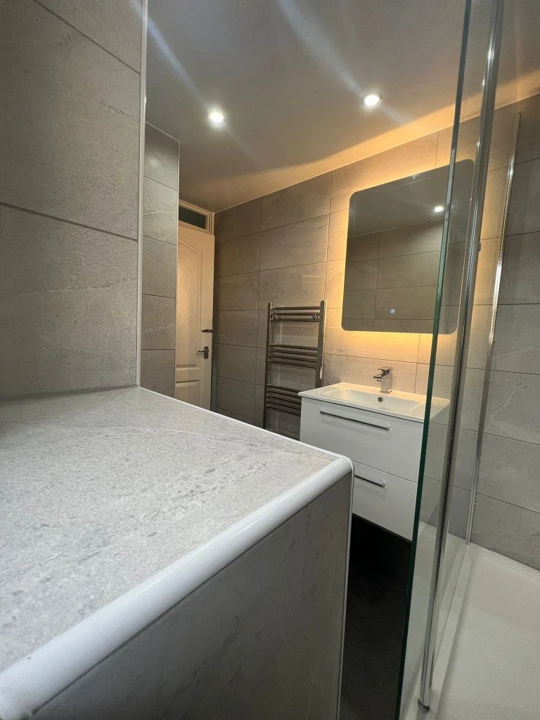 Modern bathroom with gray tiled walls, a white vanity with a sink, a mirror with backlit lighting, a heated towel rack, and a glass shower enclosure.