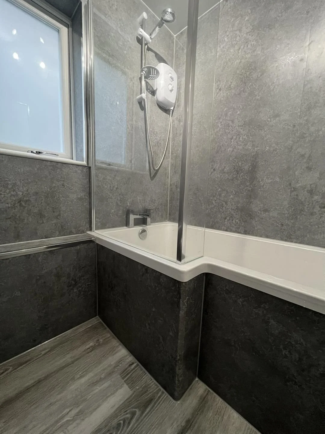 Bathroom with gray tiled walls, a bathtub with a white interior, a showerhead mounted above, a glass shower screen, a small window, and wood-patterned floor tiles.