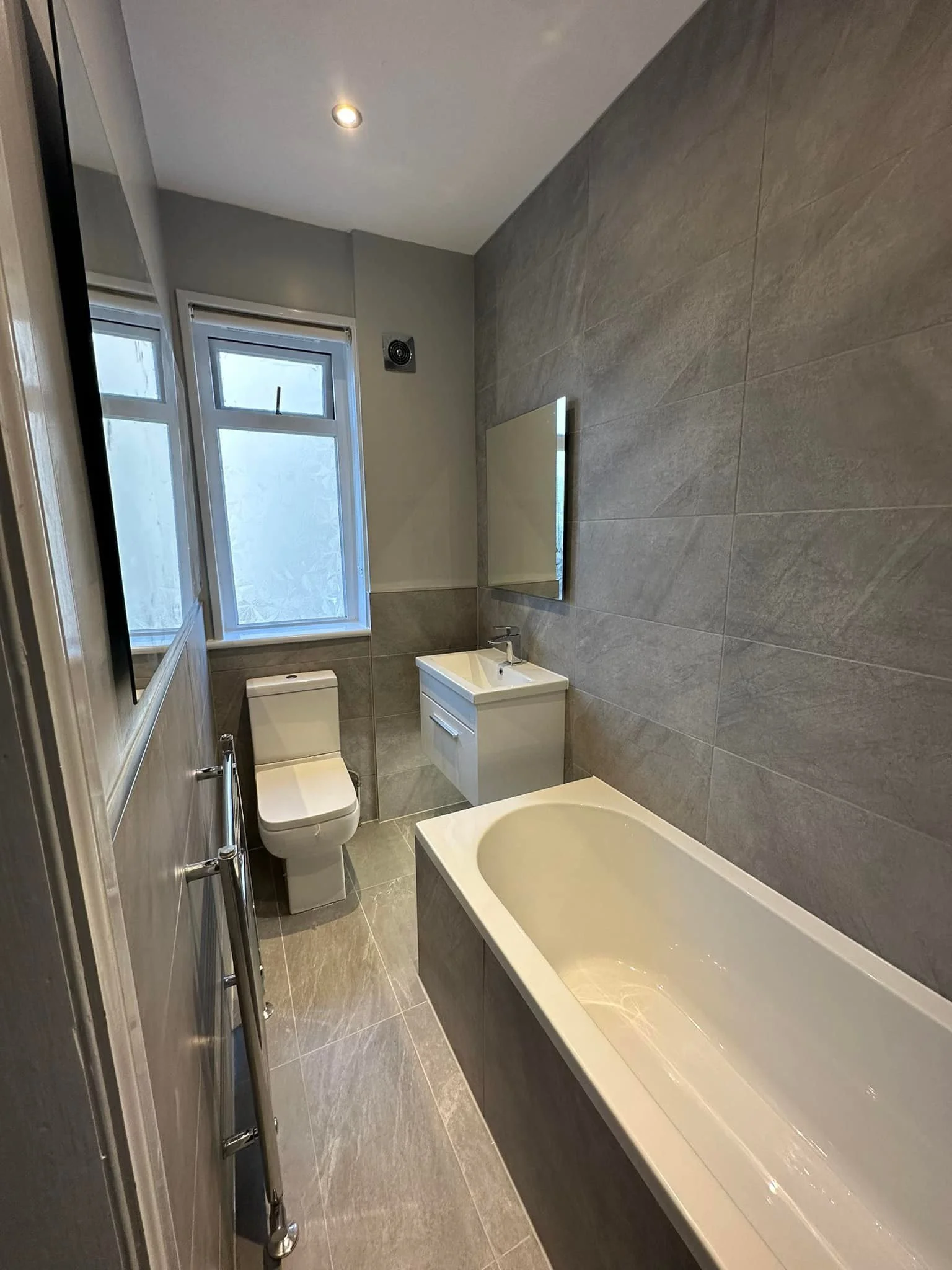 A modern bathroom with gray tiles, a white bathtub, a toilet, a small white sink with a mirror, two windows, and a ceiling light.