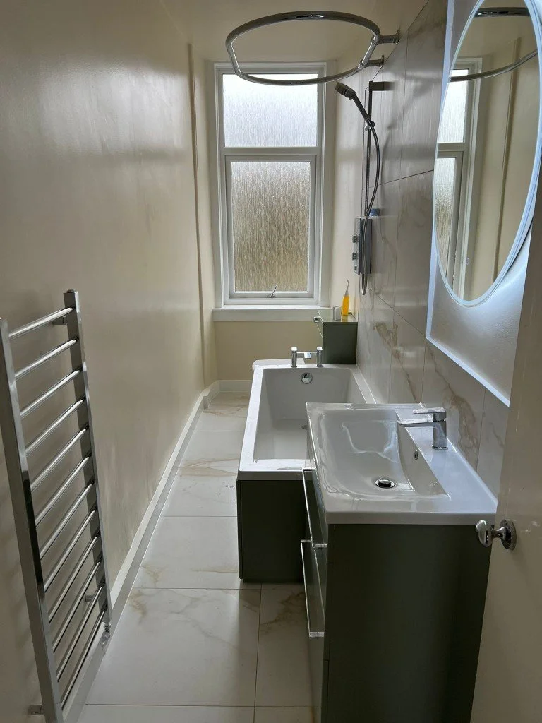 A small bathroom with a rectangular bathtub, a window with frosted glass, a wall-mounted showerhead, a white sink with a faucet, a mirror on the wall, a black soap holder, a yellow soap bottle, and a tall chrome towel radiator.