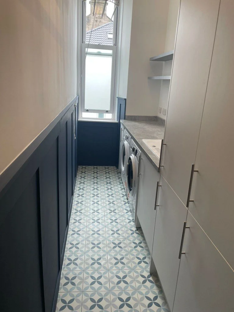 A small laundry room with a patterned tile floor, white cabinets, a countertop, washing machine, and a window at the back.