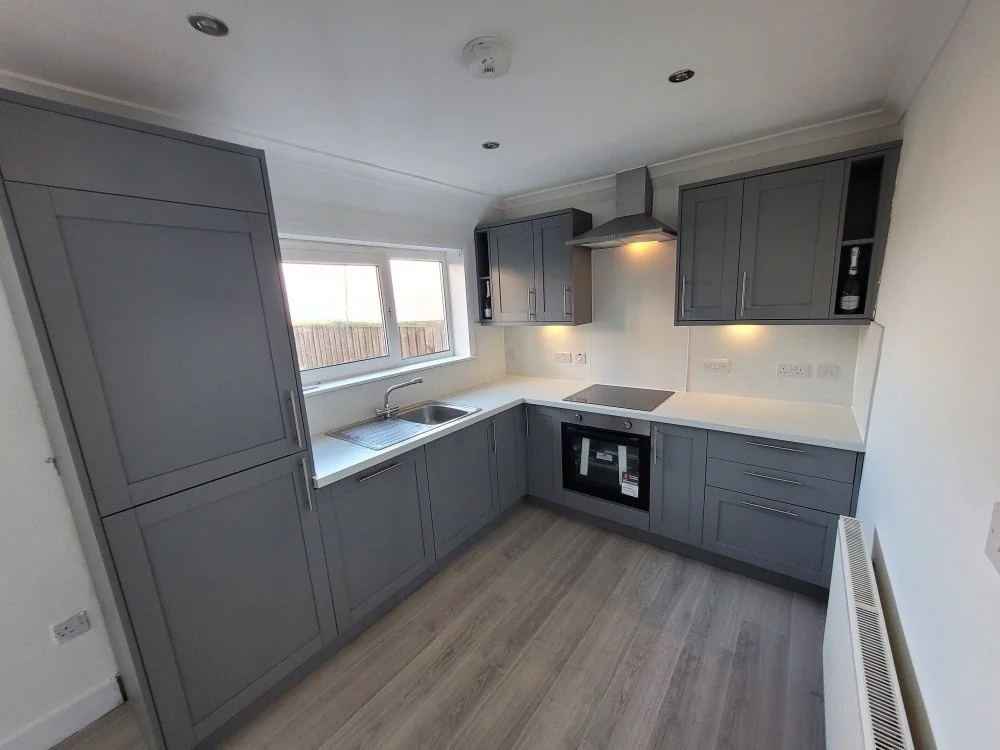 Modern kitchen with gray cabinets, white countertops, a stainless steel oven, induction cooktop, range hood, and a window above the sink.