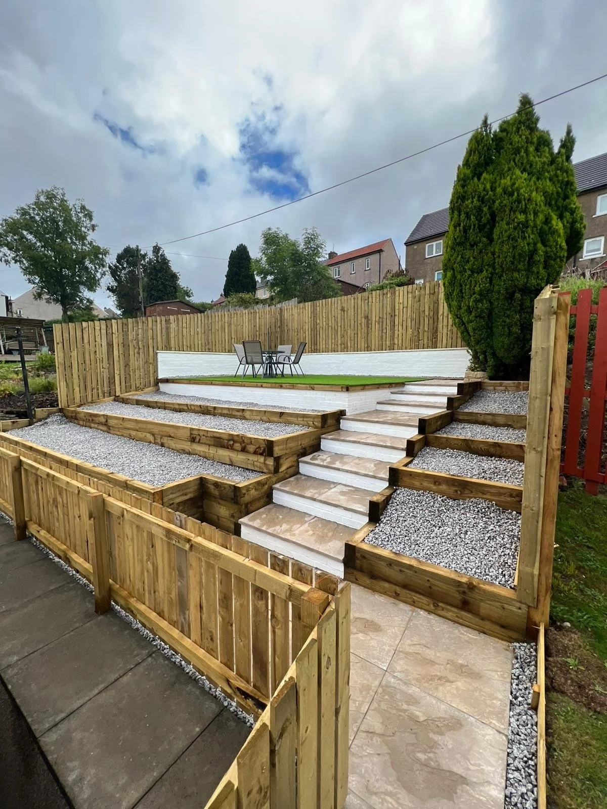 A multi-level outdoor garden with wooden fencing, stone stairs, gravel accents, a small patio with patio furniture, and green lawns and trees in the background.
