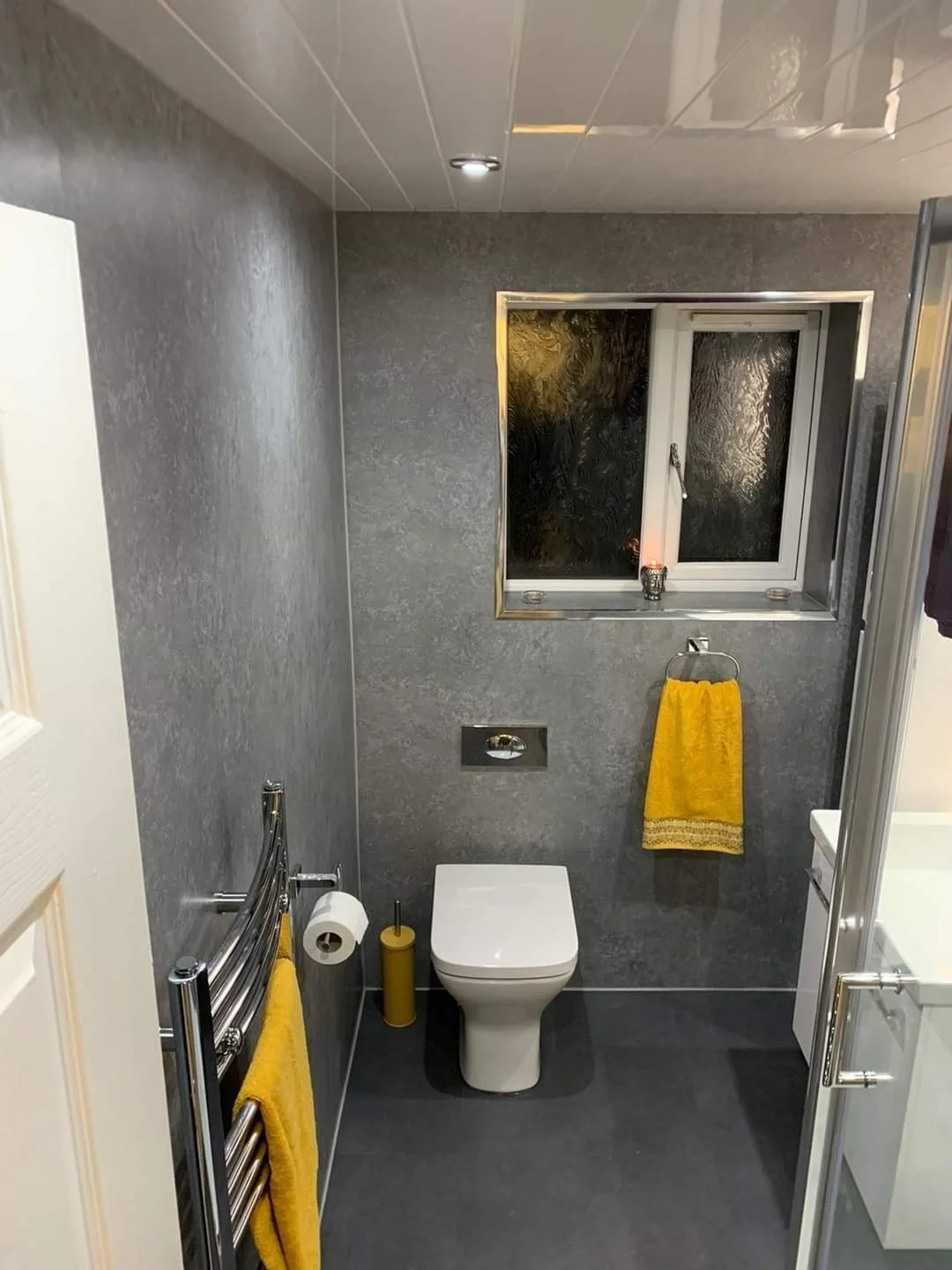Small modern bathroom with gray textured walls and floor, a white toilet, a window with black textured glass, a yellow towel on a chrome towel rack, a yellow toilet brush, and a small candle on the windowsill.