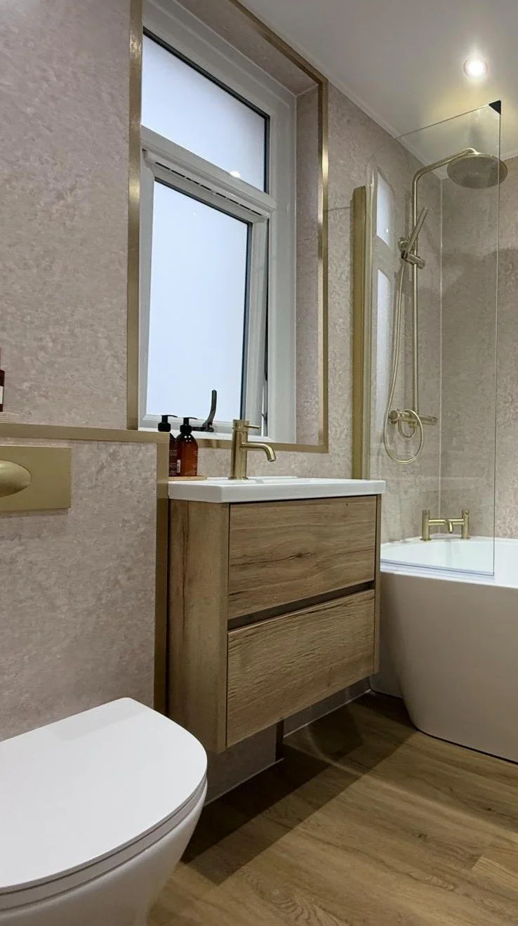 A modern bathroom with a frosted window, a wooden vanity with a sink and gold faucet, a toilet, and a bathtub with a gold faucet and a glass shower screen. There is a soap dispenser on the window sill.