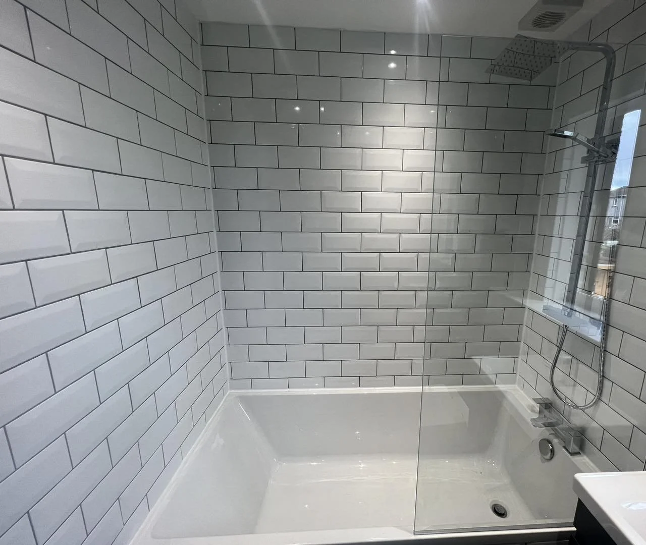 Modern shower with white subway tile walls, a large overhead showerhead, and a handheld shower attached to a wall-mounted panel. The shower is enclosed with a glass partition and has a white bathtub.