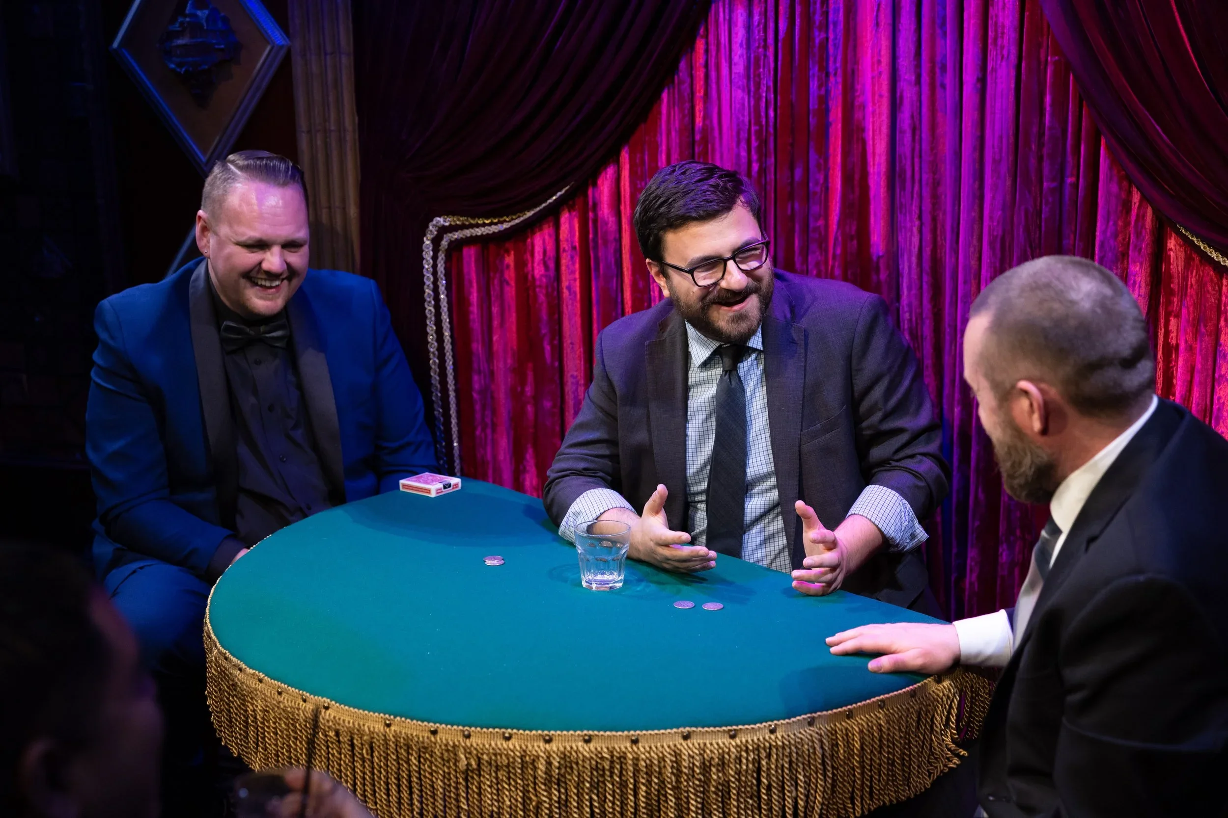 Three men sitting at a green felt card table with gold fringe, engaged in a conversation, in front of red velvet curtains. One man is smiling, another is gesturing with his hands, and the third man is listening. There are playing cards, poker chips, and a glass of water on the table.