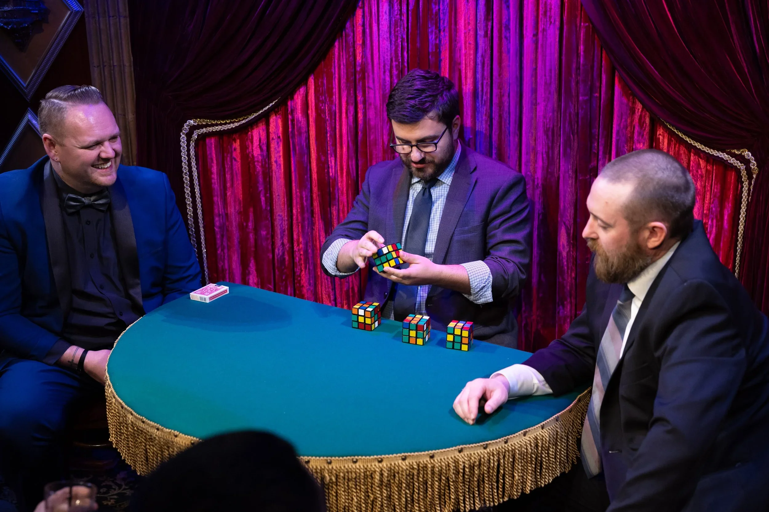 Three men sitting at a green gaming table with a golden fringe, with one man solving a Rubik's Cube, two others watching and smiling, in a room with red velvet curtains and purple lighting.