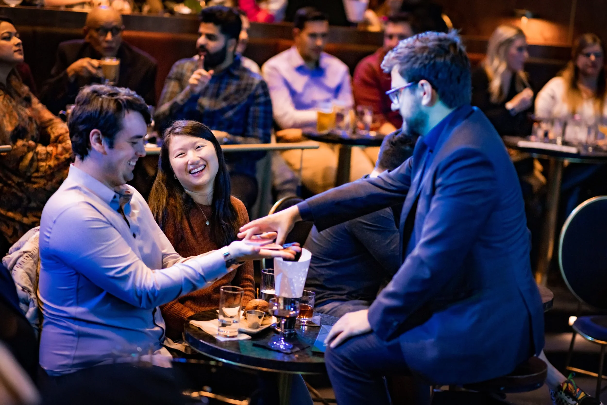 A group of people enjoying drinks and conversation at a lively social gathering or comedy show in a dimly lit venue.