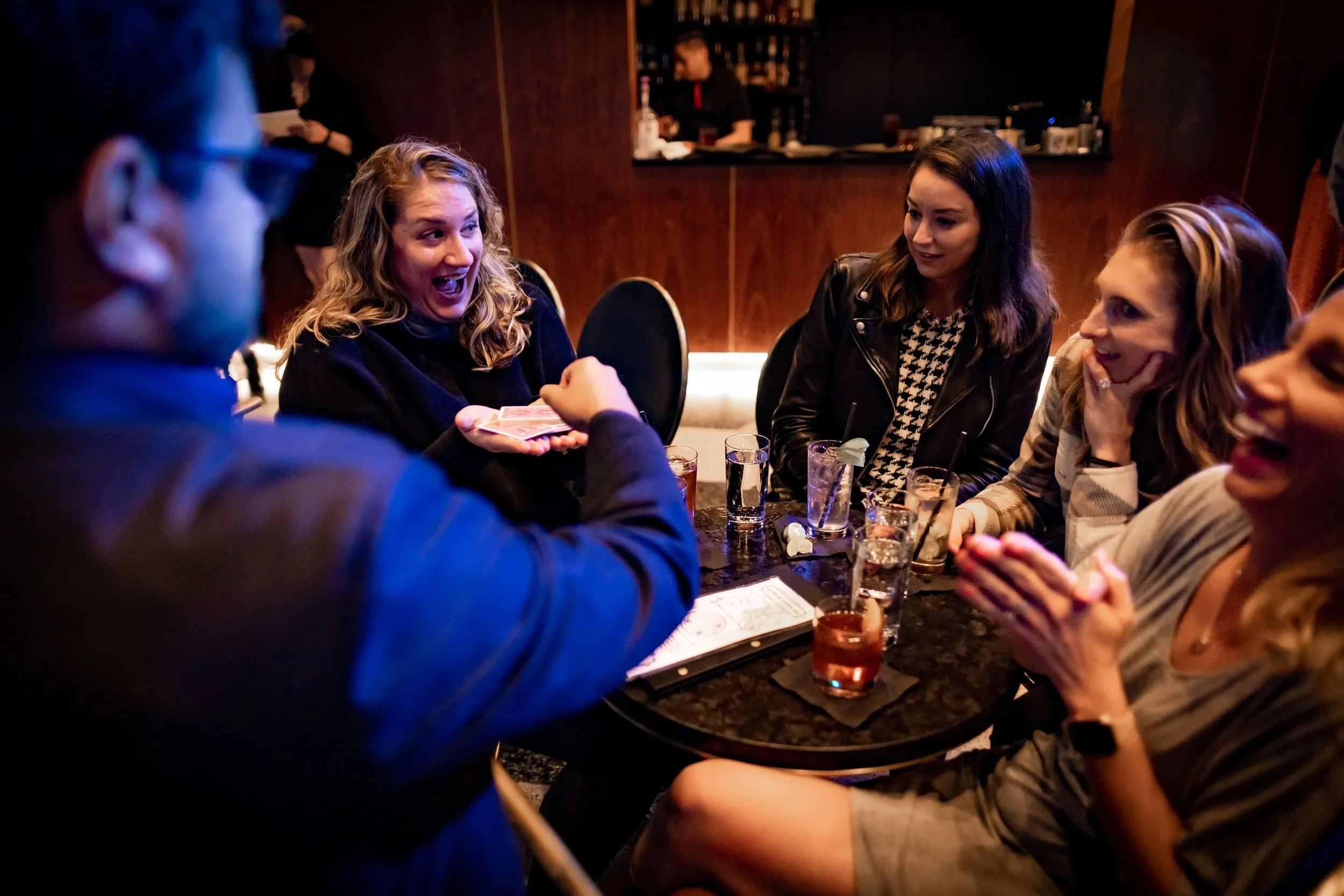 A group of five women sitting around a bar table in a dimly lit bar, engaging in a lively conversation, with one woman showing a deck of cards, others smiling and laughing, with drinks on the table.