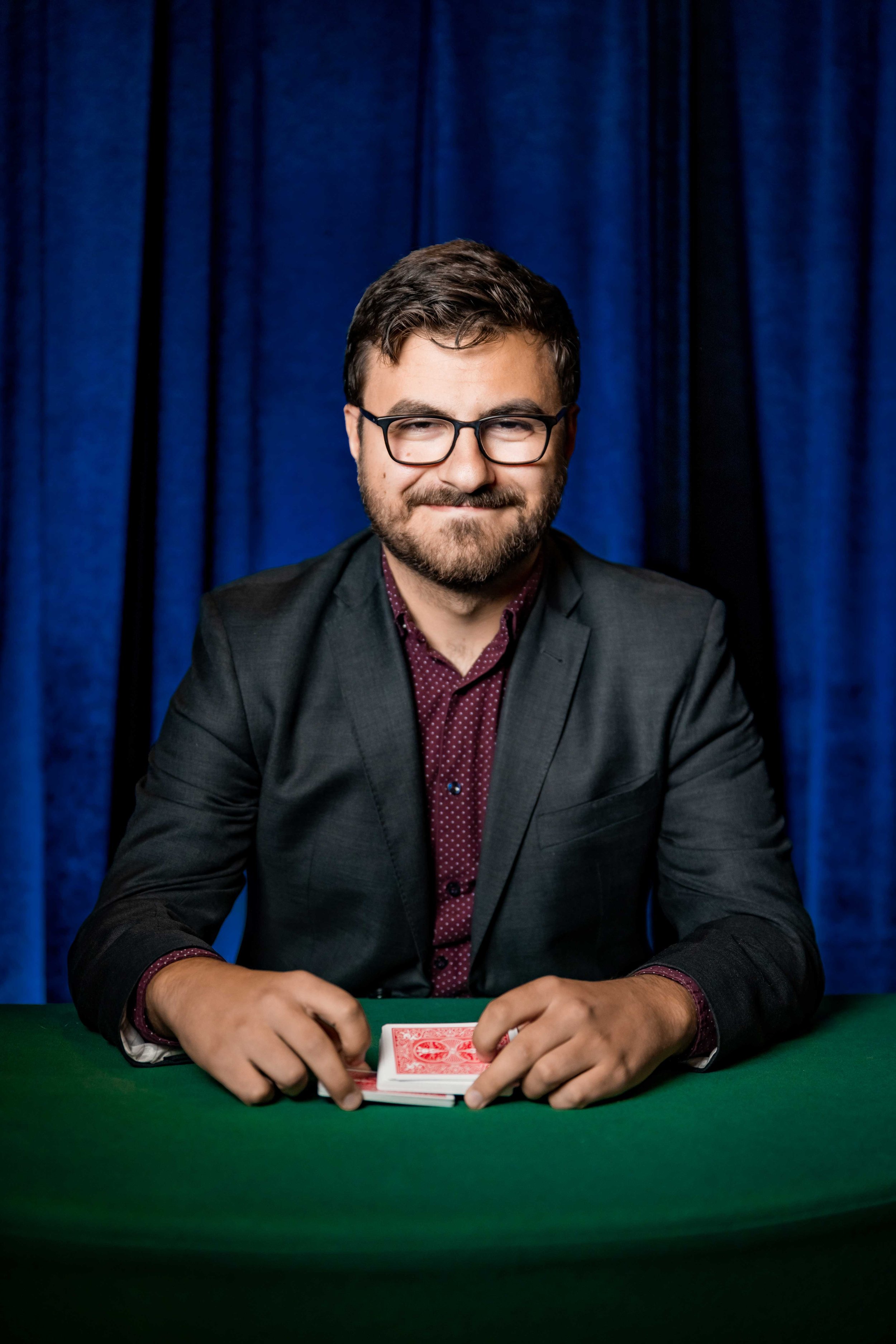A man in glasses and a black blazer seated at a green table with playing cards, smiling, with a blue curtain background.
