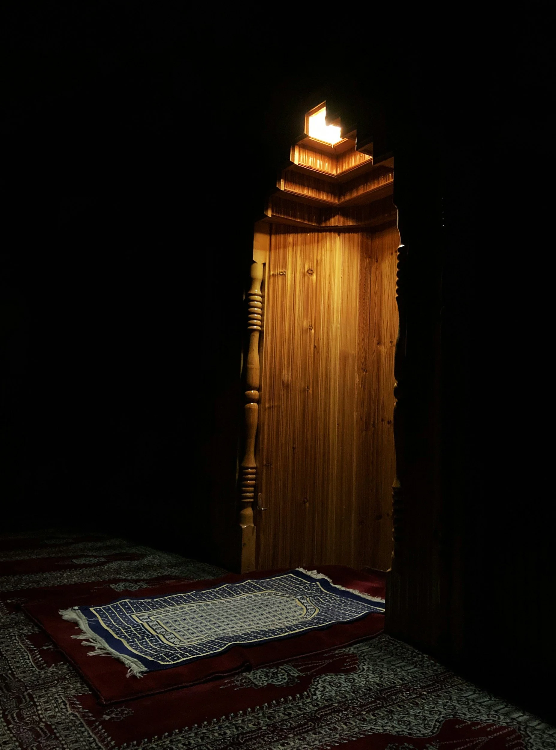 A dimly lit room with a wooden door illuminated from behind, a prayer rug on the floor, and a wooden lattice above the door opening to a bright light.