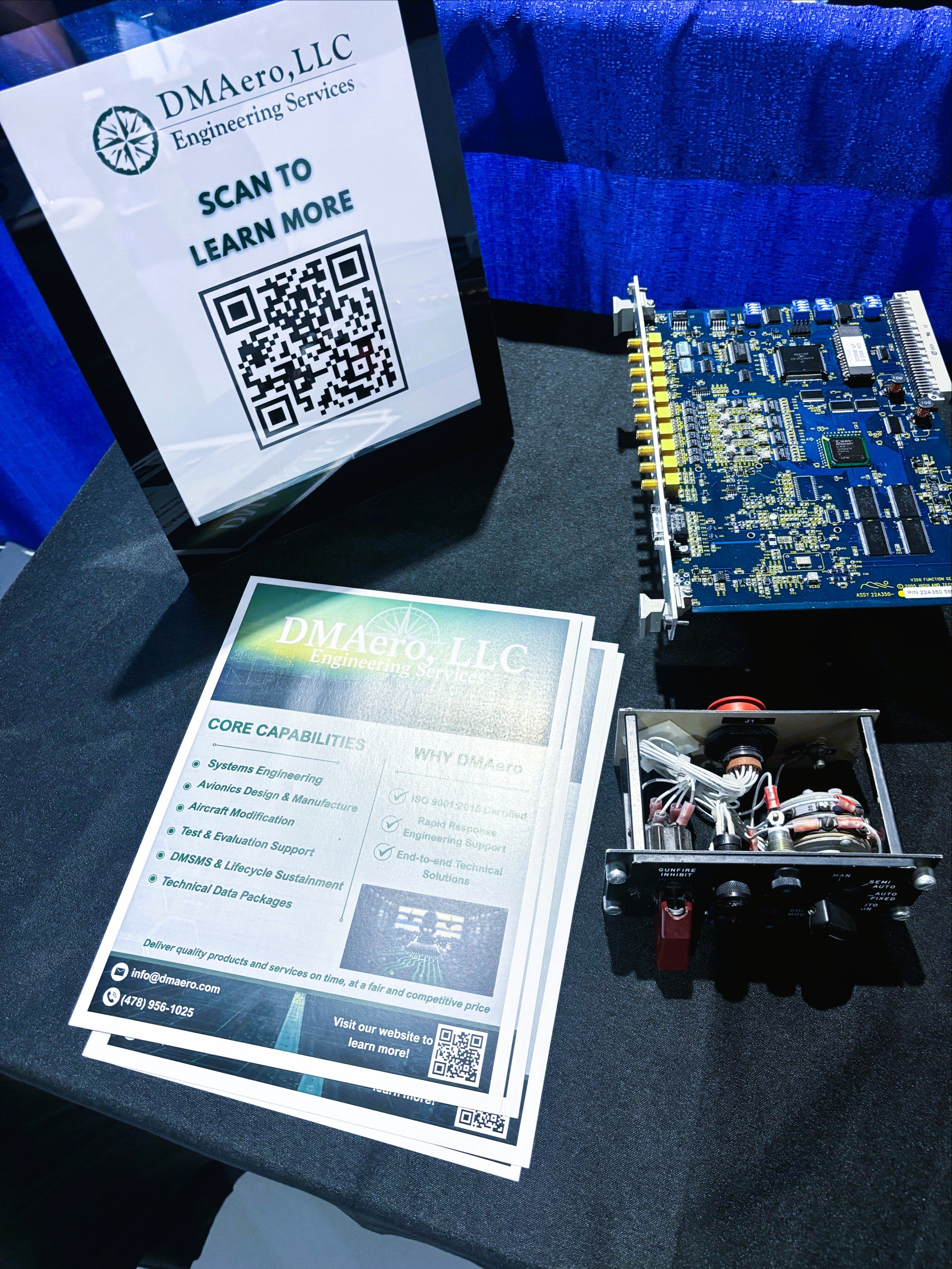 Display booth of DMAero LLC Engineering Services featuring a QR code sign, a printed brochure, a blue circuit board, and electronic components on a black table.