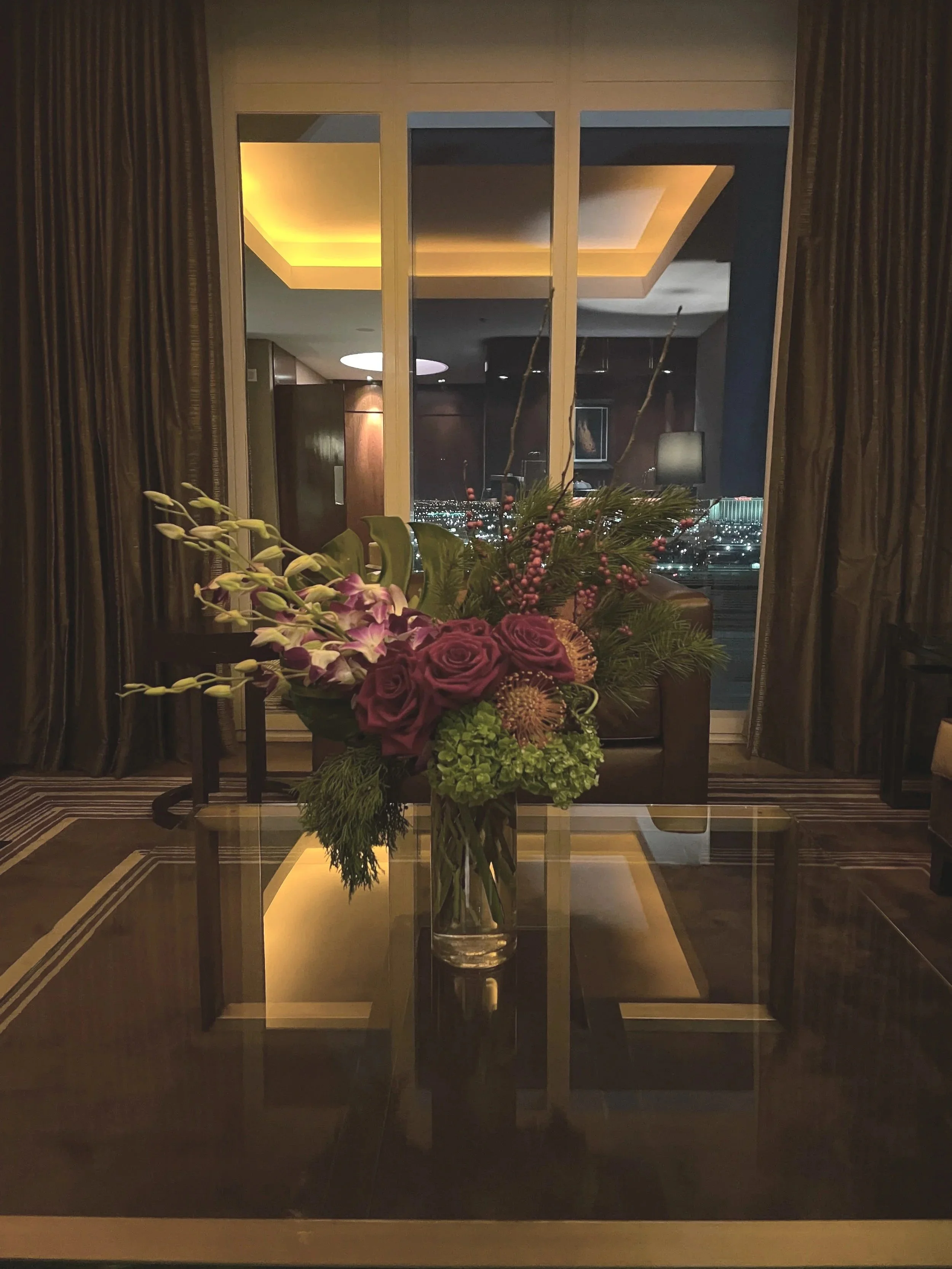 A flower arrangement featuring purple roses, green hydrangeas, pink berries, purple orchids, and greenery in a tall glass vase on a glass table.