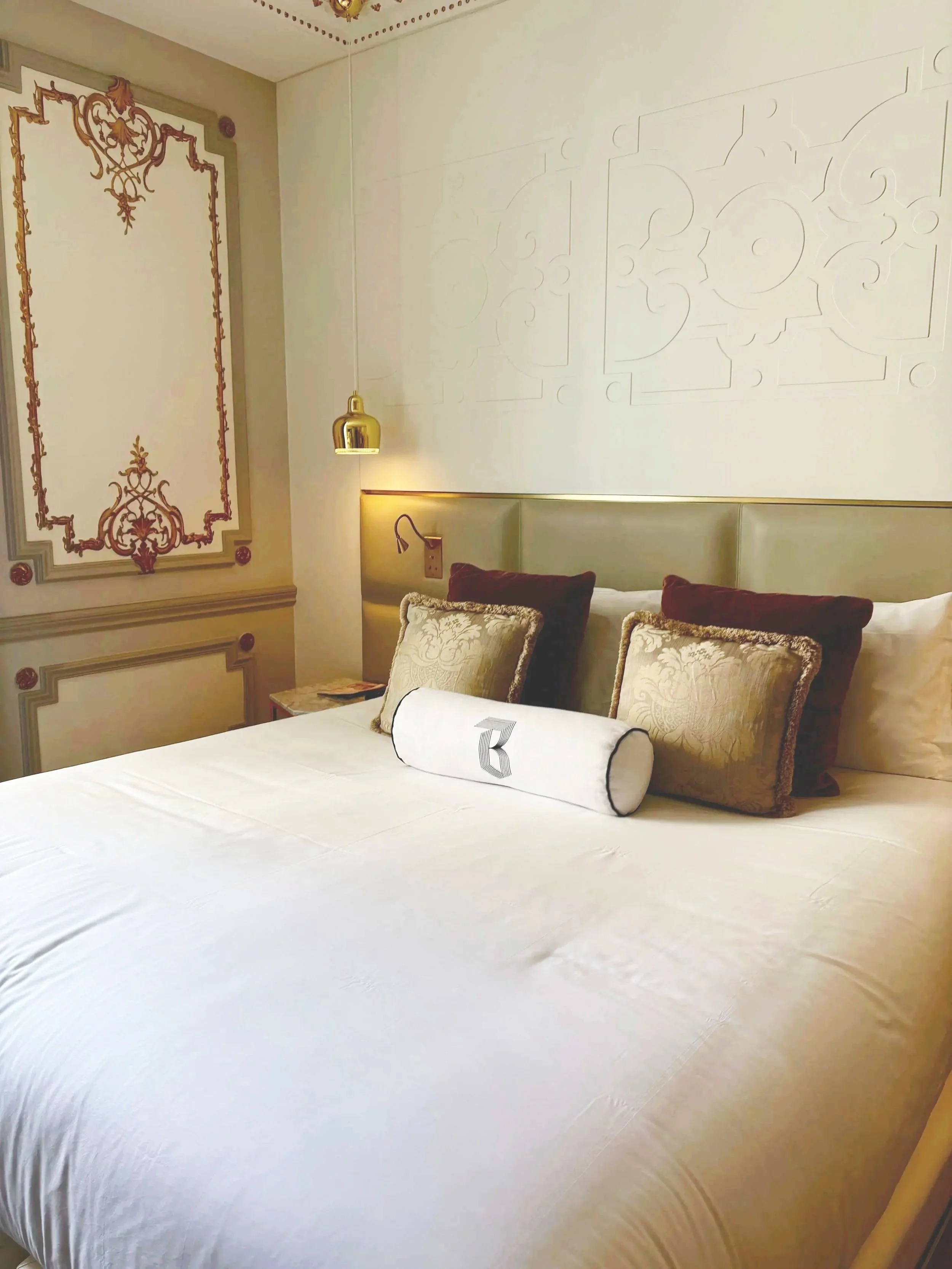 A neatly made hotel bed with decorative pillows and a cylindrical bolster pillow, in a room with ornate wall designs, a hanging gold lamp, and a bedside table.