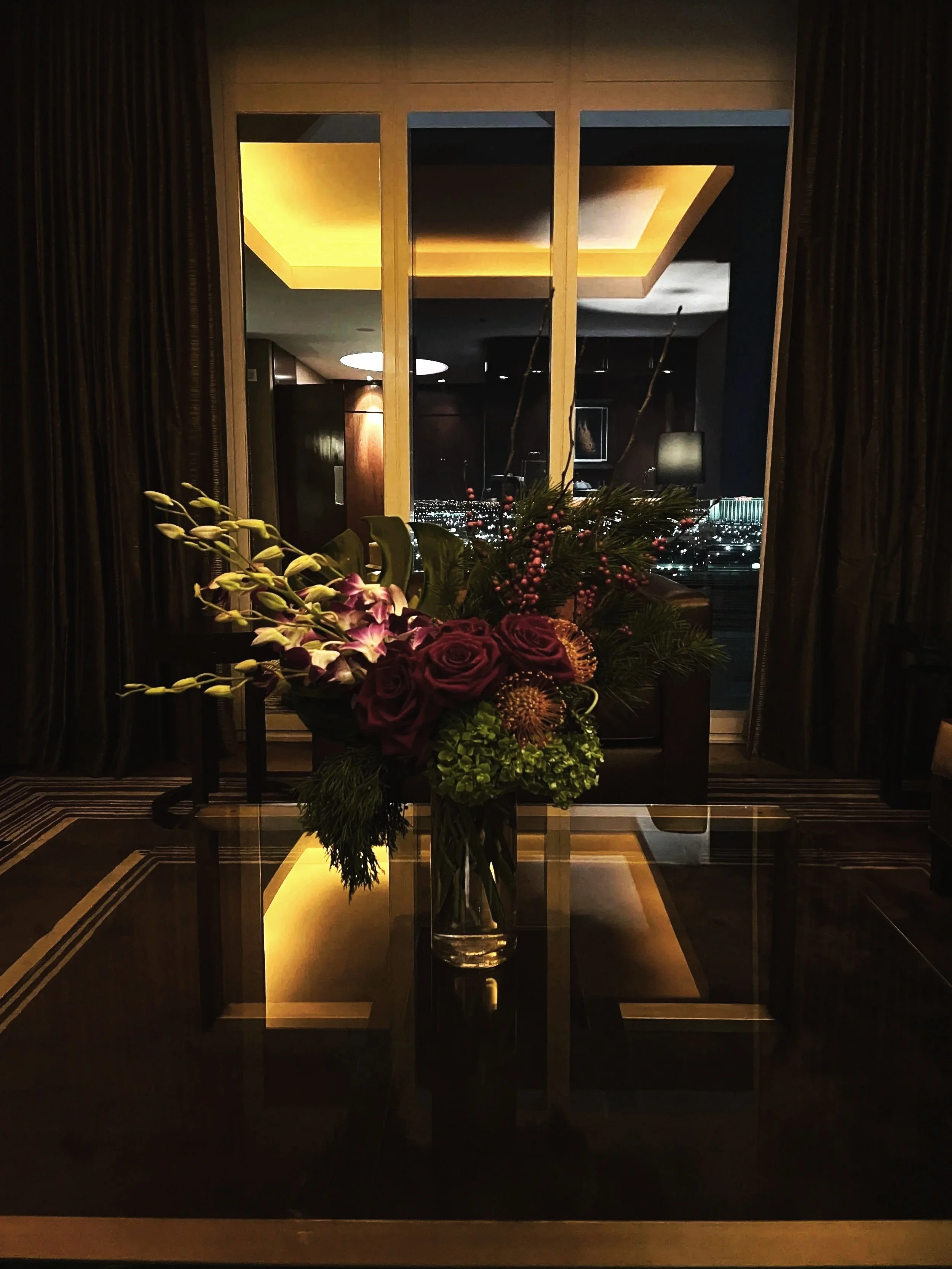A flower arrangement featuring purple roses, green hydrangeas, pink berries, purple orchids, and greenery in a tall glass vase on a glass table.