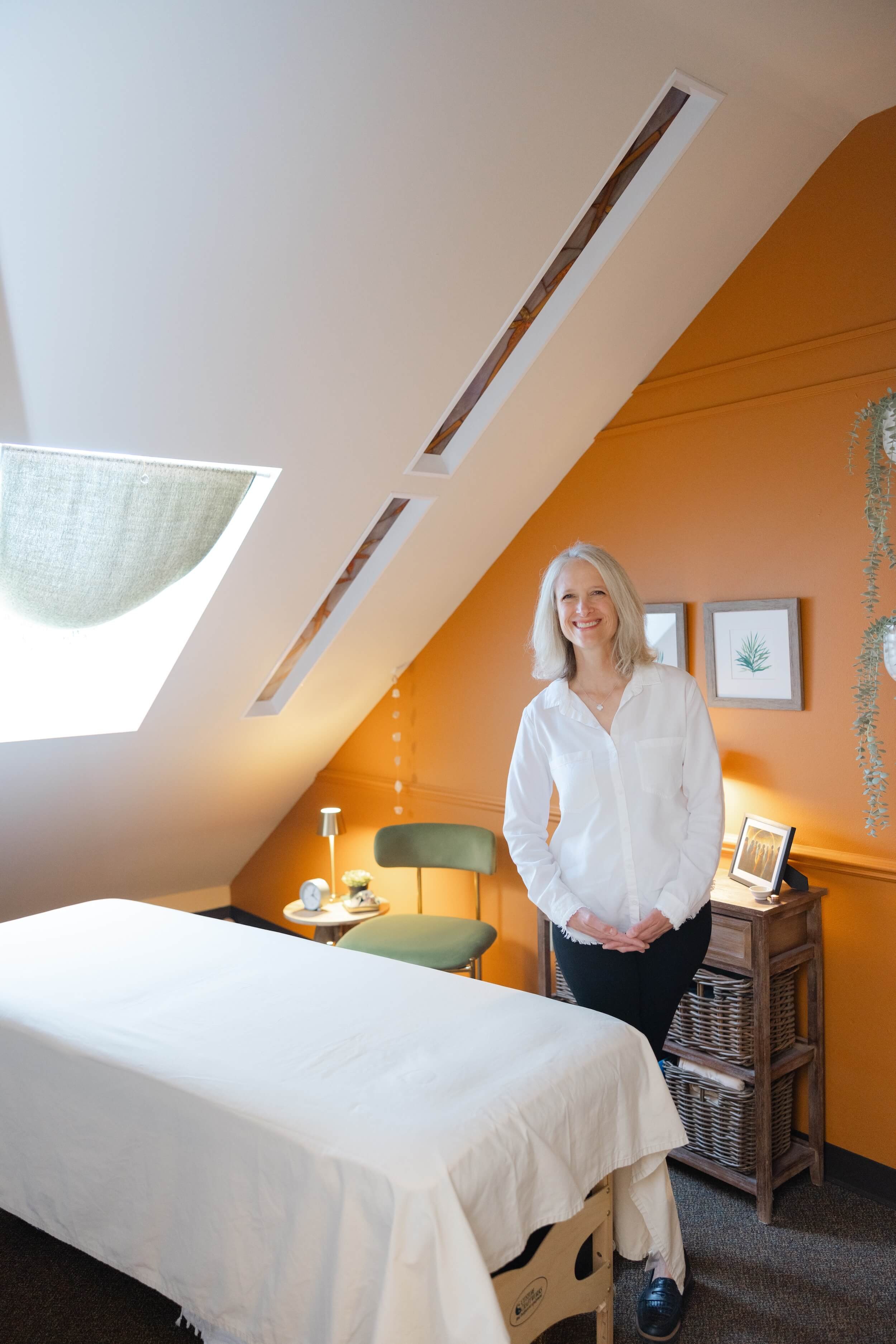 Eleanor Nowacki standing beside a massage table in a cozy room with orange walls and framed artwork, sunlight coming through a skylight, small side table with plants and notes, and a green chair.
