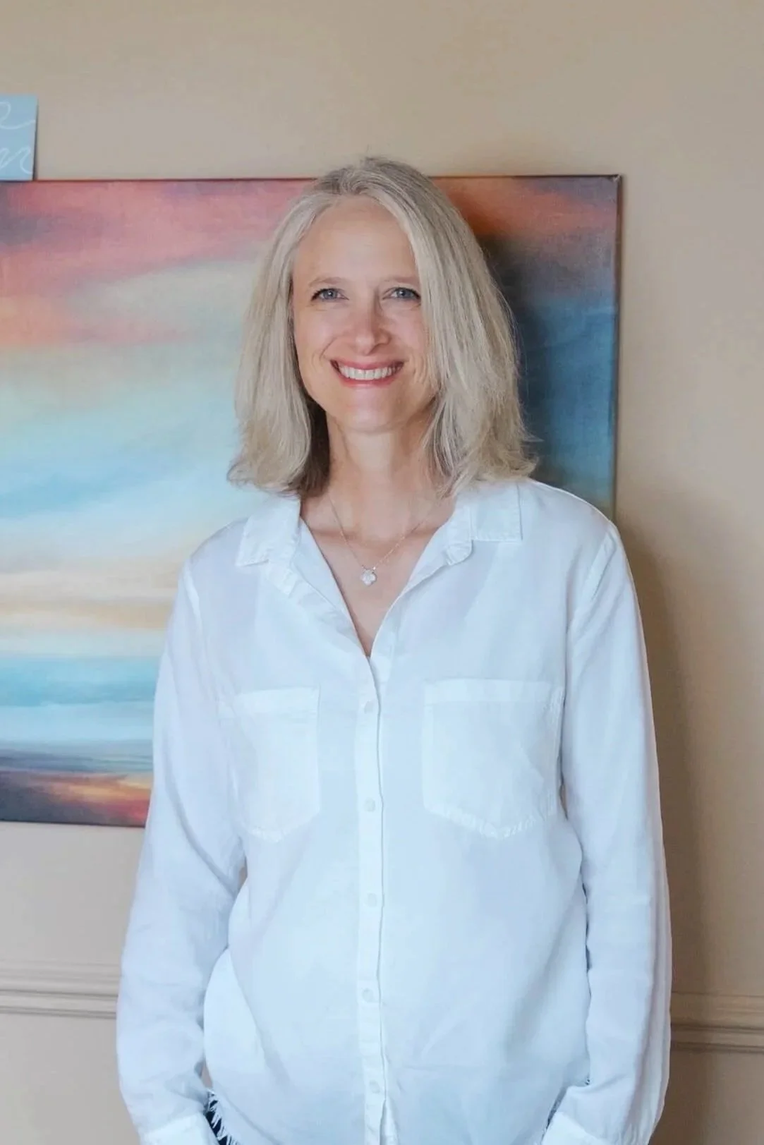 Eleanor Nowacki, a woman with blonde hair, wearing a white shirt, smiling, standing in front of a colorful abstract painting on a beige wall.