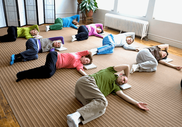 Group of adults practicing Feldenkrais Awareness Through Movement® on a brown mat indoors, lying in various relaxed poses.