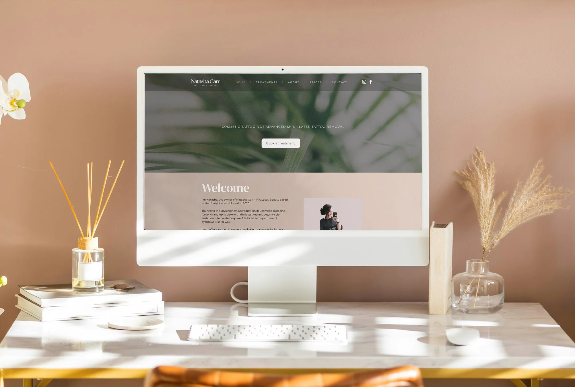 A computer monitor on a white desk displays a website for Natasha Carr, a beauty and tattooing service provider. The desk has a stack of white books, a small glass jar with reed diffusers, a white plate, a keyboard, and a white mouse. To the right of the monitor, there is a glass vase with dried pampas grass and a wooden decorative object. The background wall is painted in a soft beige color.