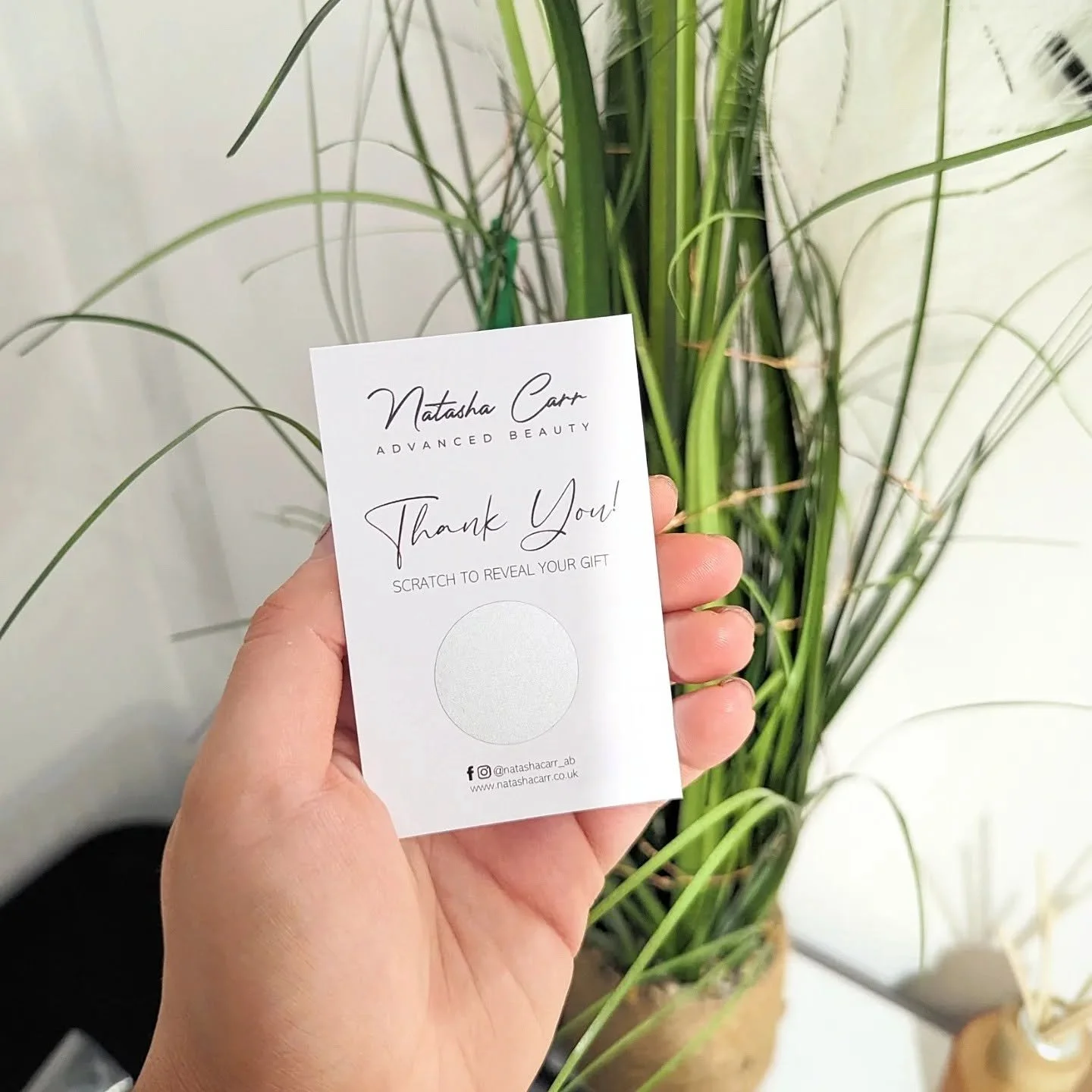 Person holding a white card with text that says 'Natasha Carr, Advanced Beauty, Thank You! Scratch to Reveal Your Gift,' in front of a tall green houseplant with long, slender leaves.