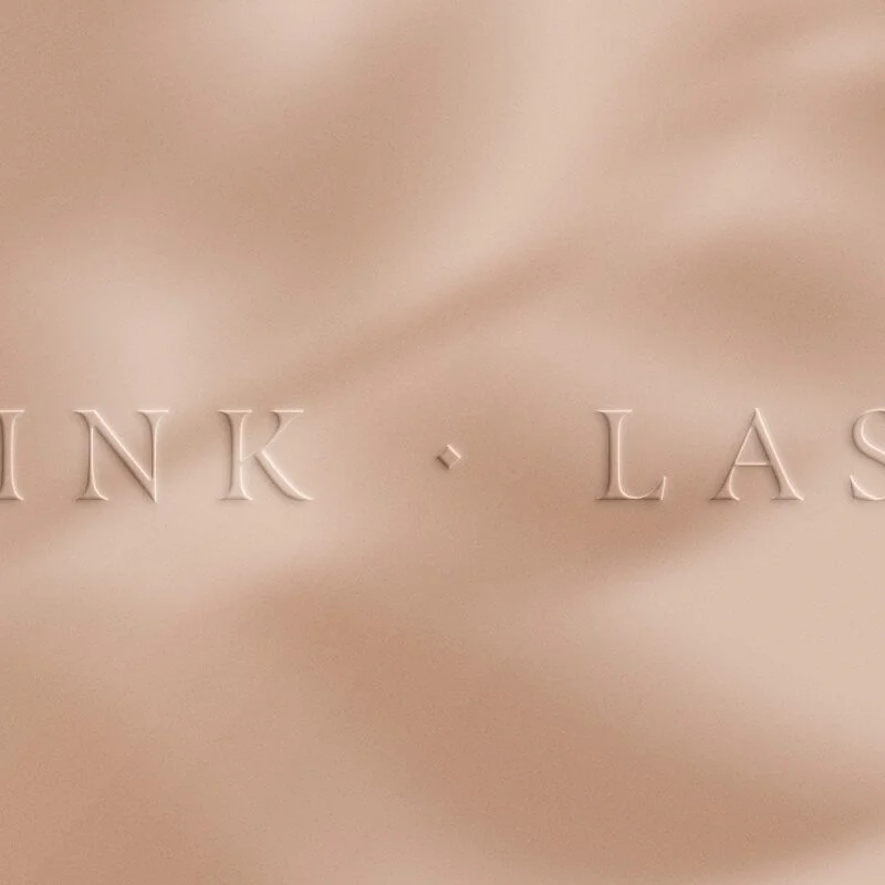 A close-up, debossed 'INK · LASER' logo for Ink Laser, set against a warm sand-colored background with soft, ethereal leaf shadows.