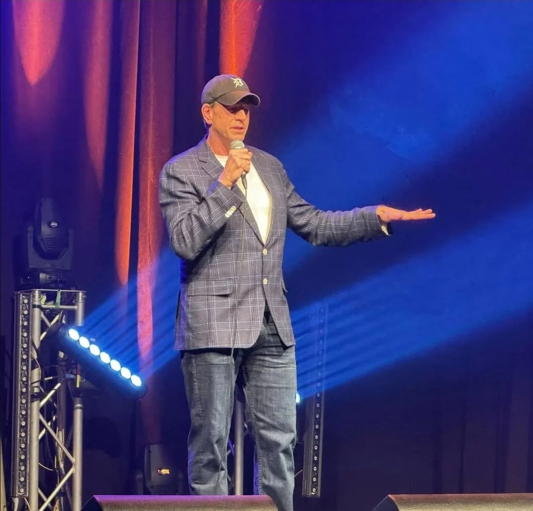 A man wearing a black cap, sunglasses, a checkered blazer, a white shirt, and jeans, holding a microphone and performing on stage with blue and orange lighting and stage equipment.