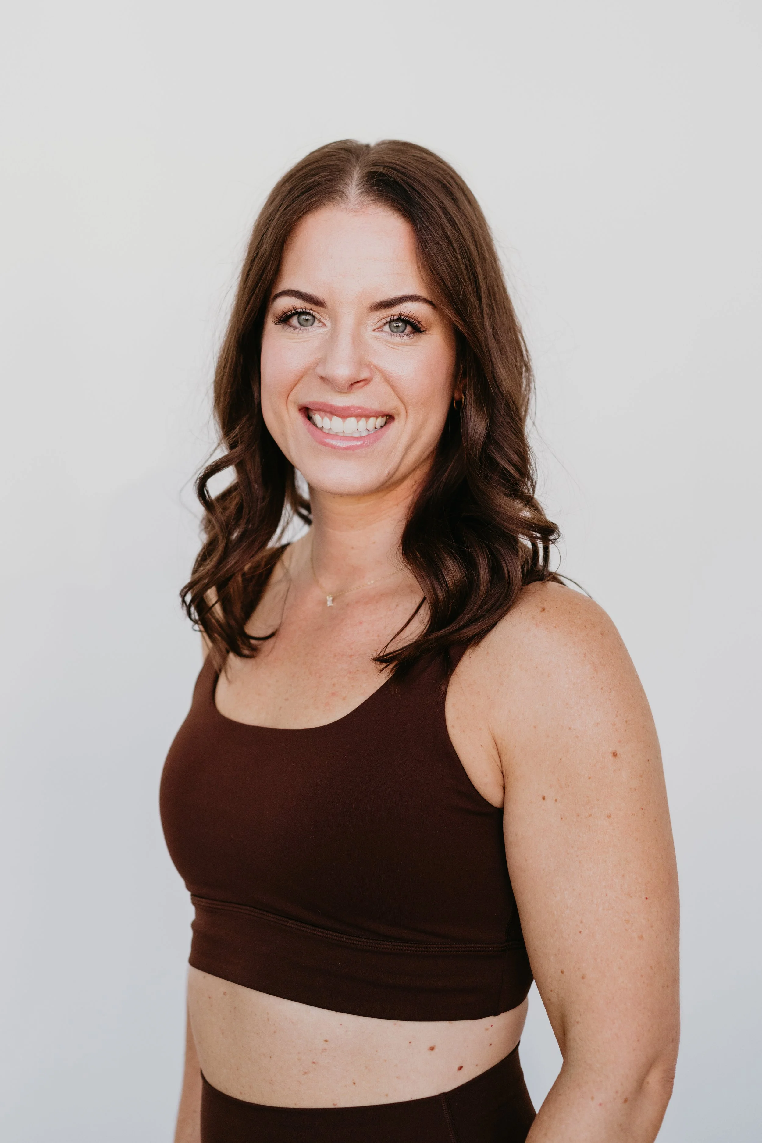 A woman with long wavy brown hair, wearing a dark brown sports bra, smiling against a plain white background.