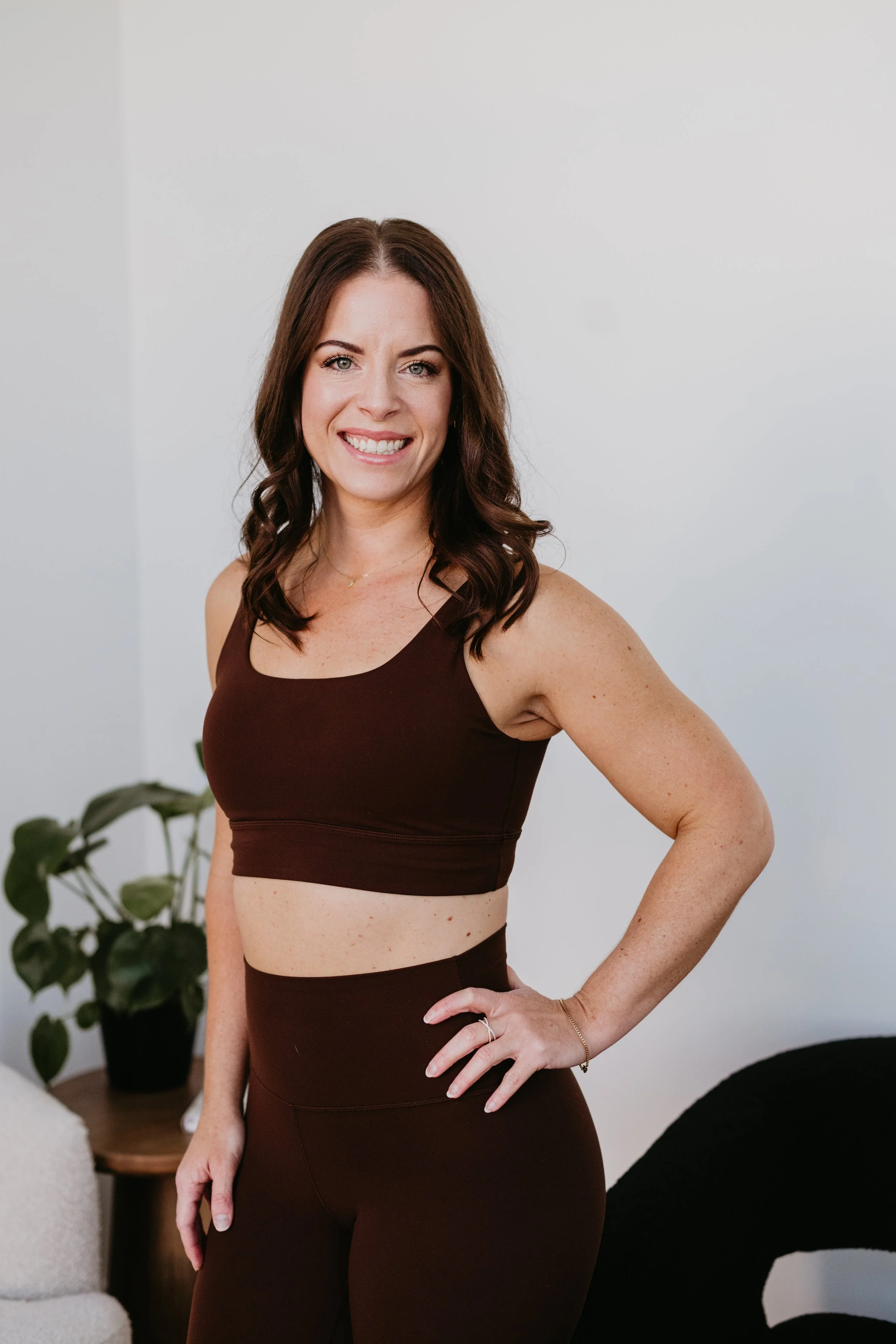 A woman with long brown hair in loose curls, wearing a brown sports bra and matching high-waisted leggings, stands in a minimalistic room with a plant in the background. She is smiling and has one hand on her hip.