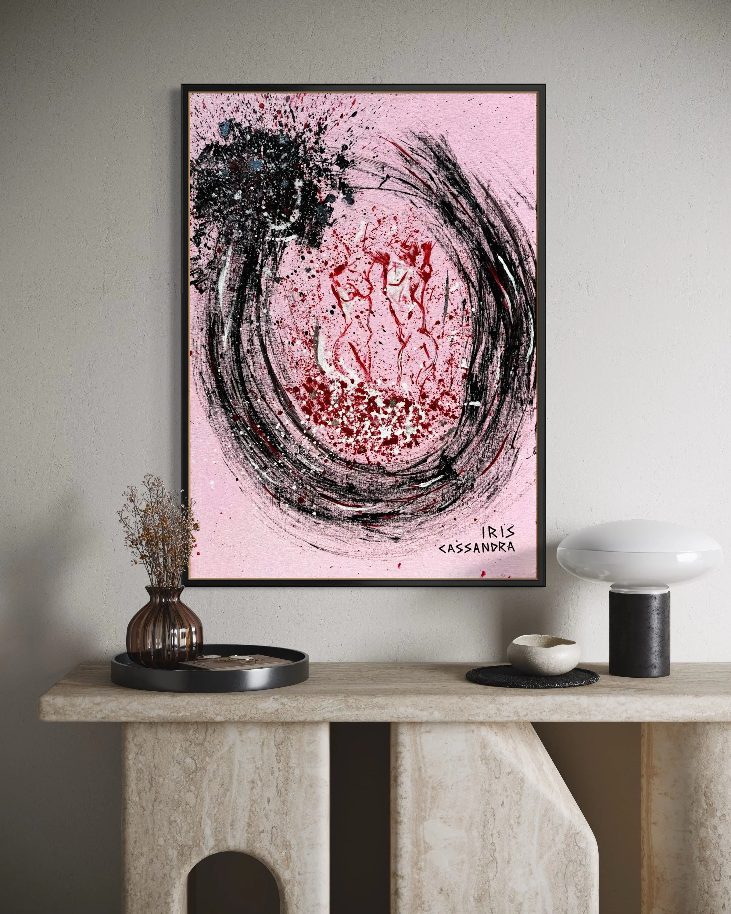 Abstract painting with pink background, black and red splashes, and swirling brushstrokes, signed by Iris Cassandra, hanging on a wall above a beige marble console table with decorative items.