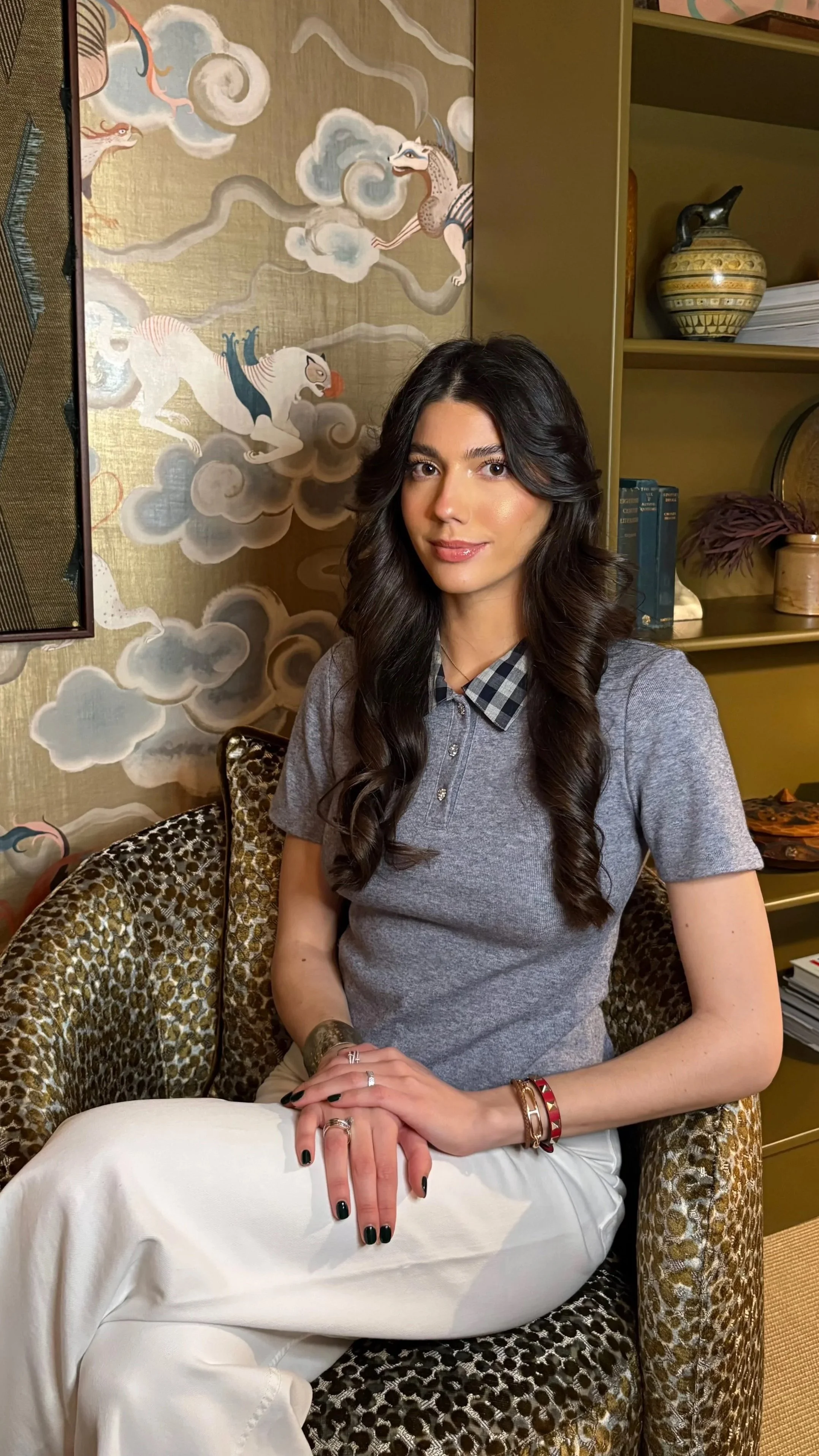 A young woman with long wavy dark hair and light skin sitting on chair with animal print fabric; wearing a gray shirt with a checkered collar, white pants, and jewelry including rings, bracelets, and a watch. Behind her is a decorative wall with a fantasy-style mural of dragons and clouds, and a shelf with books, vases, and decor.