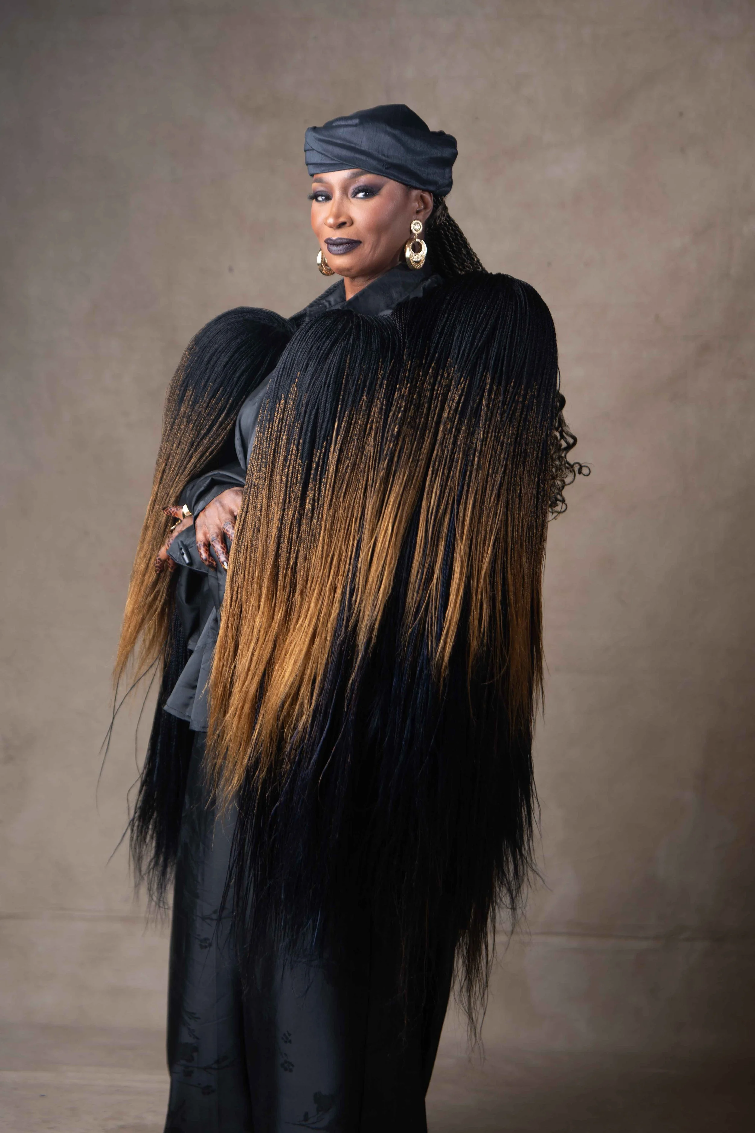 A woman with dark skin, wearing a black headwrap, dark lipstick, and bold earrings, dressed in a black outfit with long, ombré dreadlocks that transition from black to light brown, standing against a neutral background.