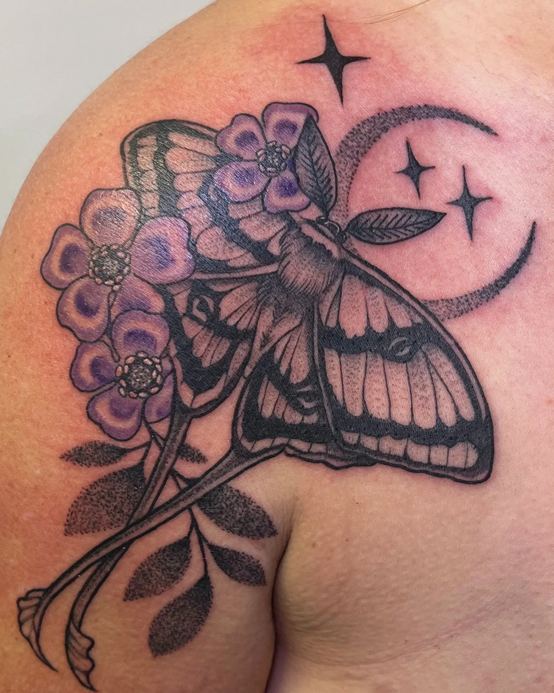 Tattoo of a butterfly on a shoulder with purple flowers, leaves, a crescent moon, and three stars.