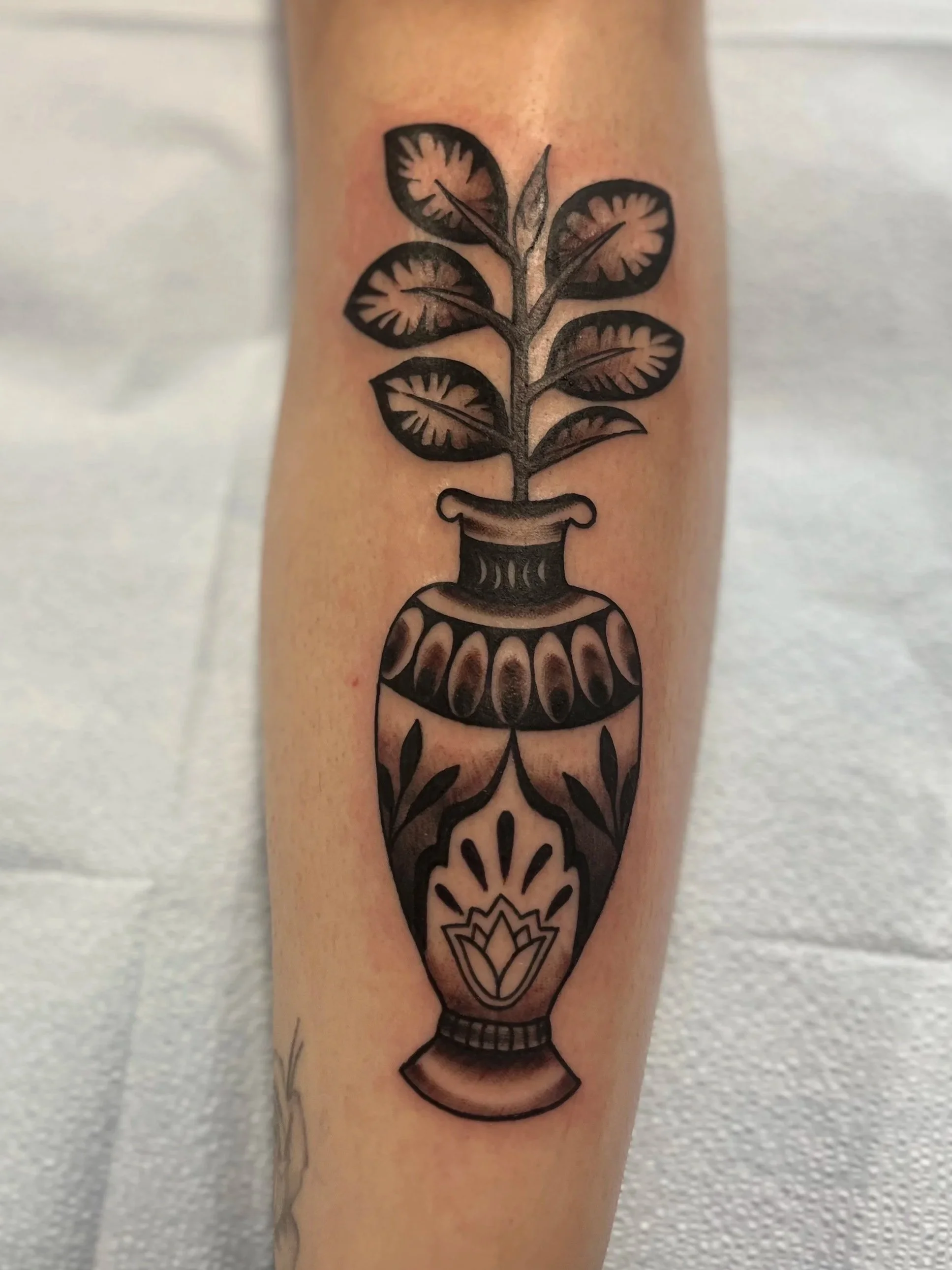 A black line tattoo of a vase with a plant growing out of it on a person's shin.