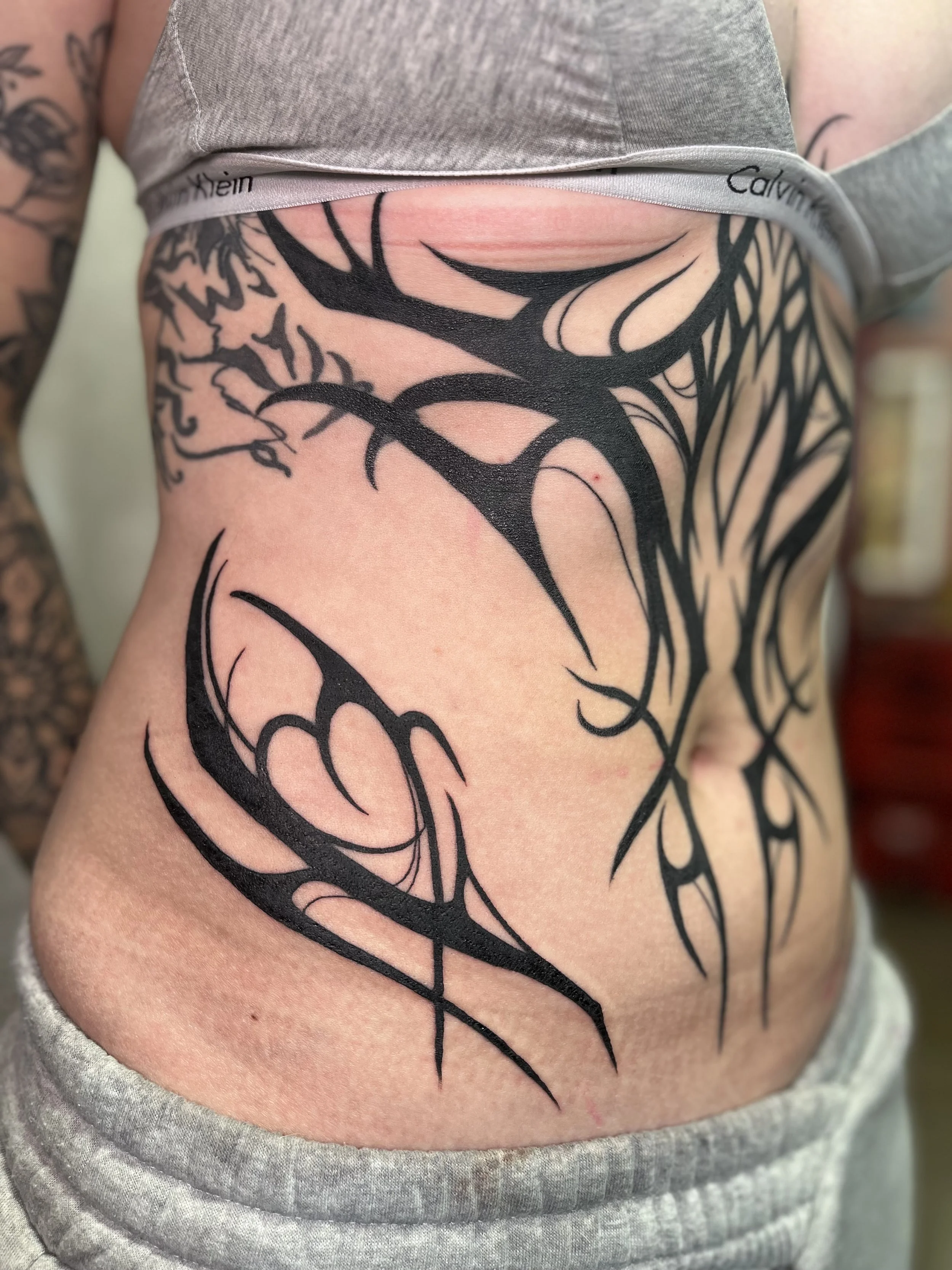 Close-up of a person's side torso with a large black new school tribal-style tattoo, cybersigilism, blackwork, incorporating butterflies and hearts