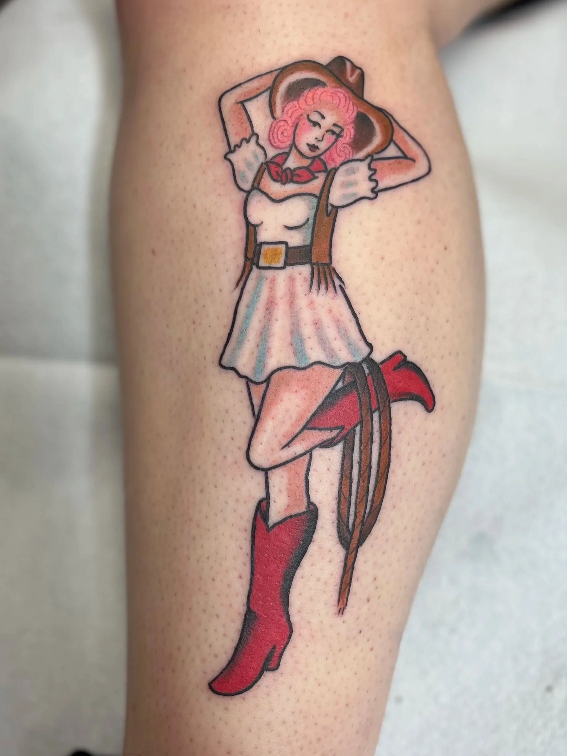 Tattoo of a burlesque cowboy woman with pink hair, wearing a cowboy hat, red cowboy boots, a short skirt, and a red neckerchief, posing with arms behind her head.