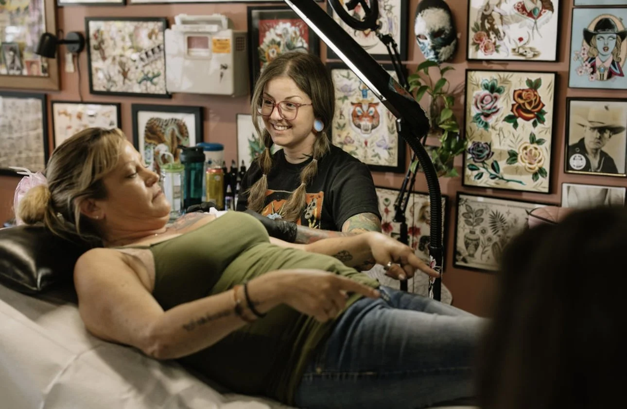 Tattoo artist tattooing a woman on her arm in a tattoo studio decorated with framed tattoo art on the walls.