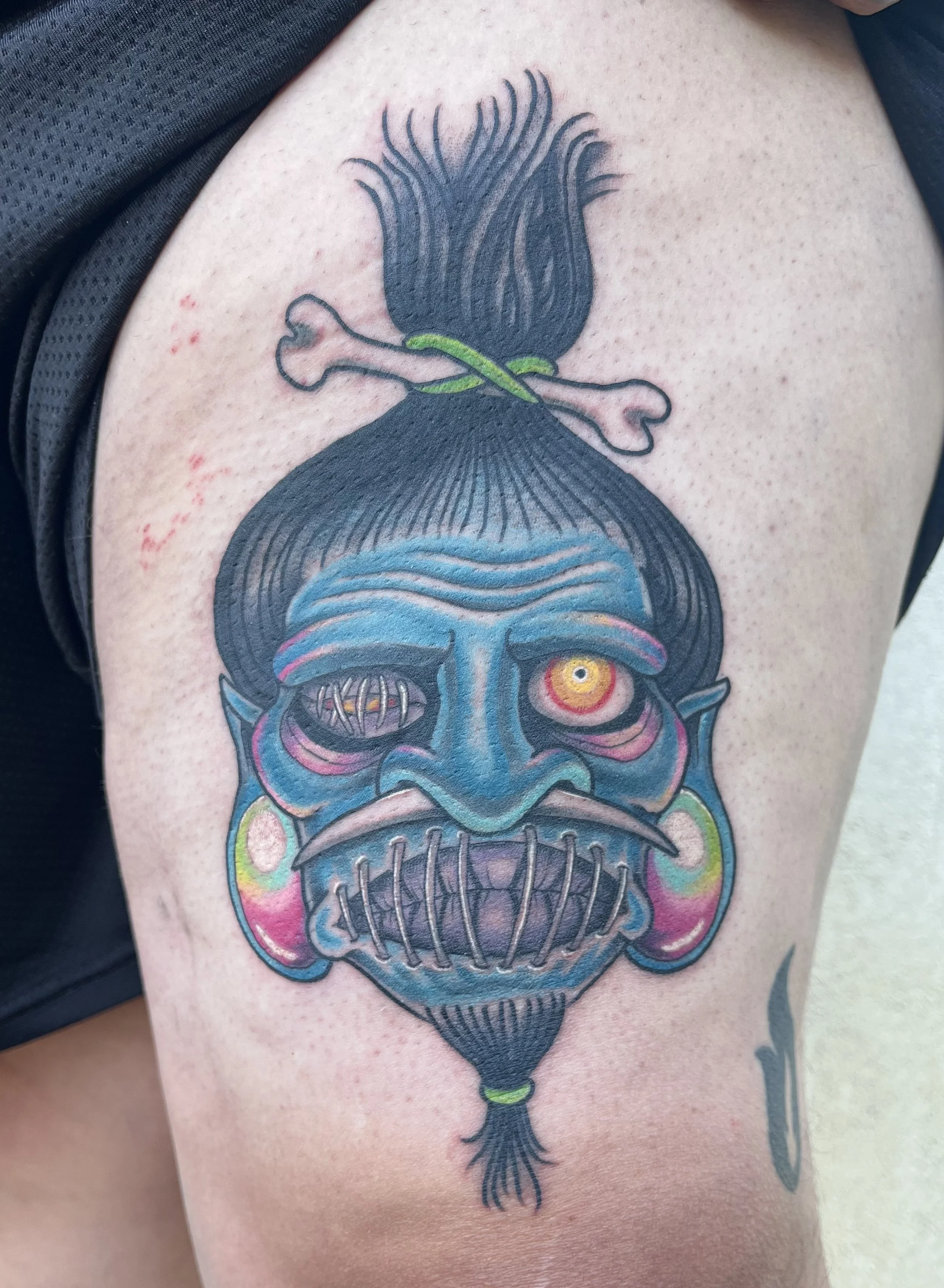 Colorful tattoo of a blue skull with one yellow eye and one closed eye, adorned with earrings and a pointy beard, with a black hair bun tied with a green strap, on a person's upper arm.